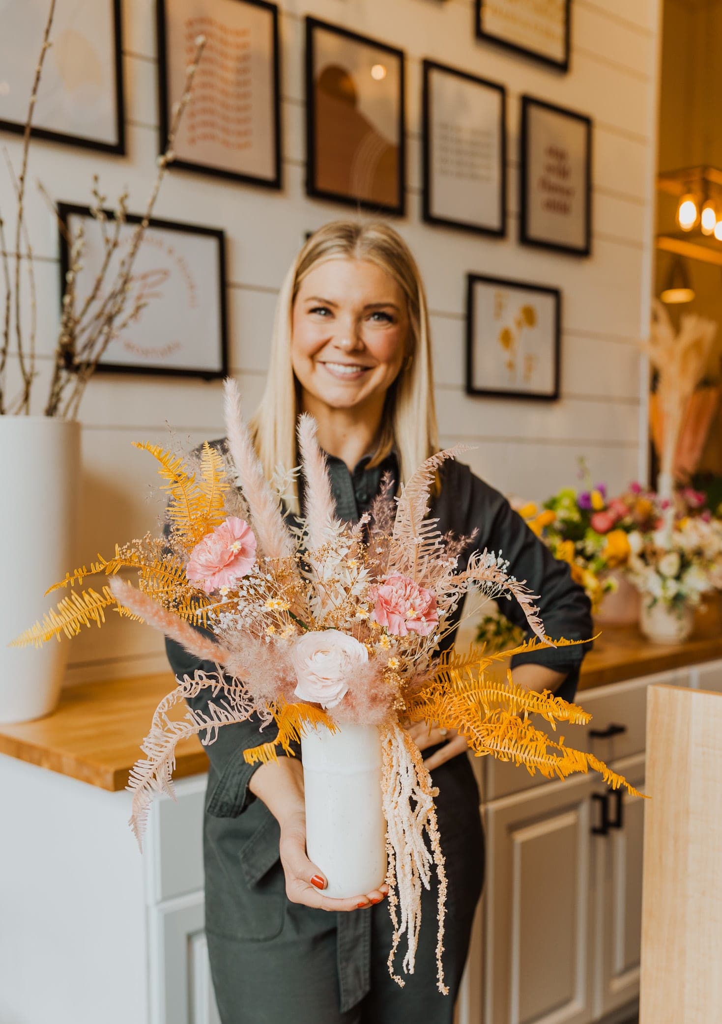 Florist holding a tall white vase with pink flowers and feathery golden dried accents in a shop.