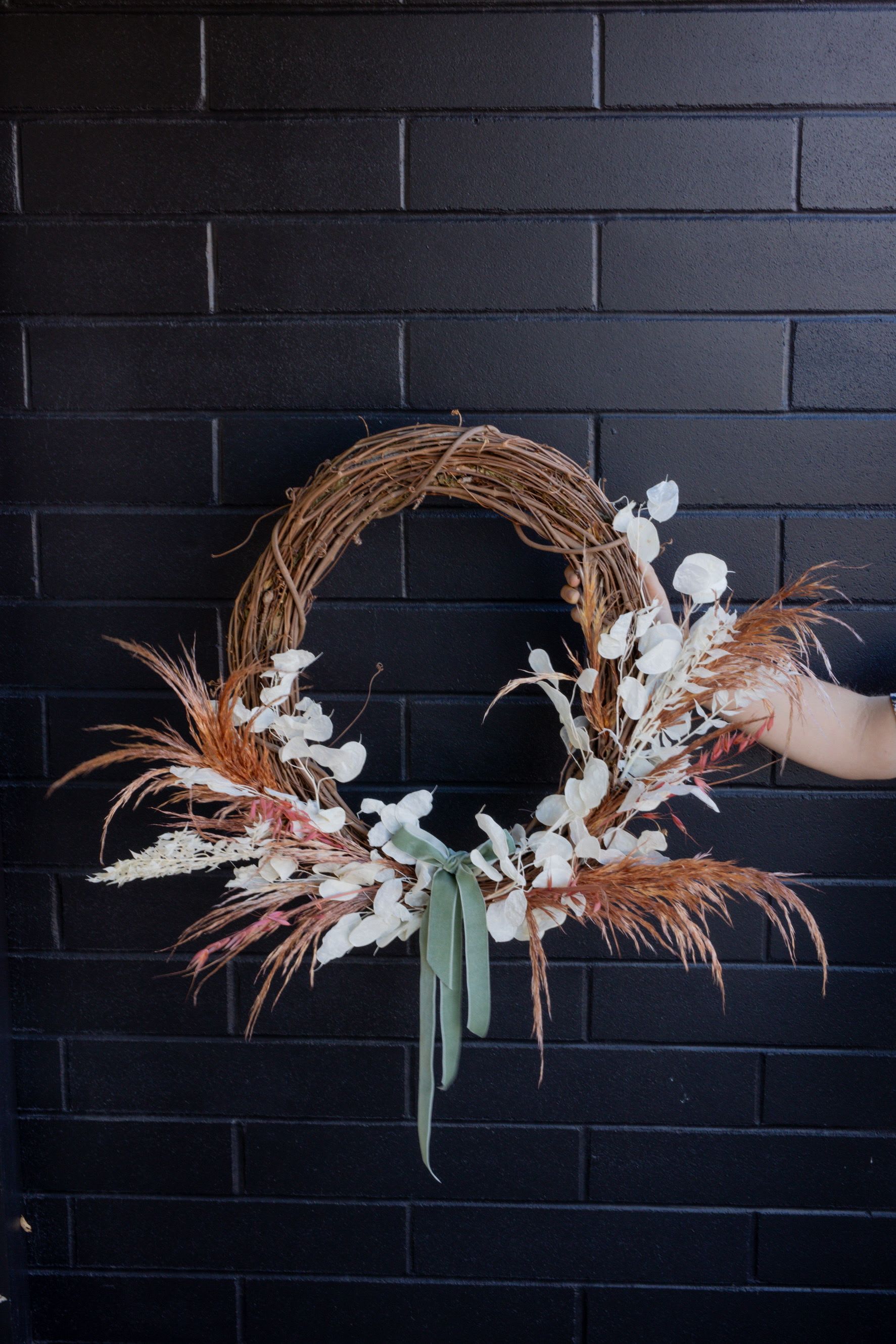 Asymmetrical dried floral wreath with white accents and sage ribbon on a grapevine base.