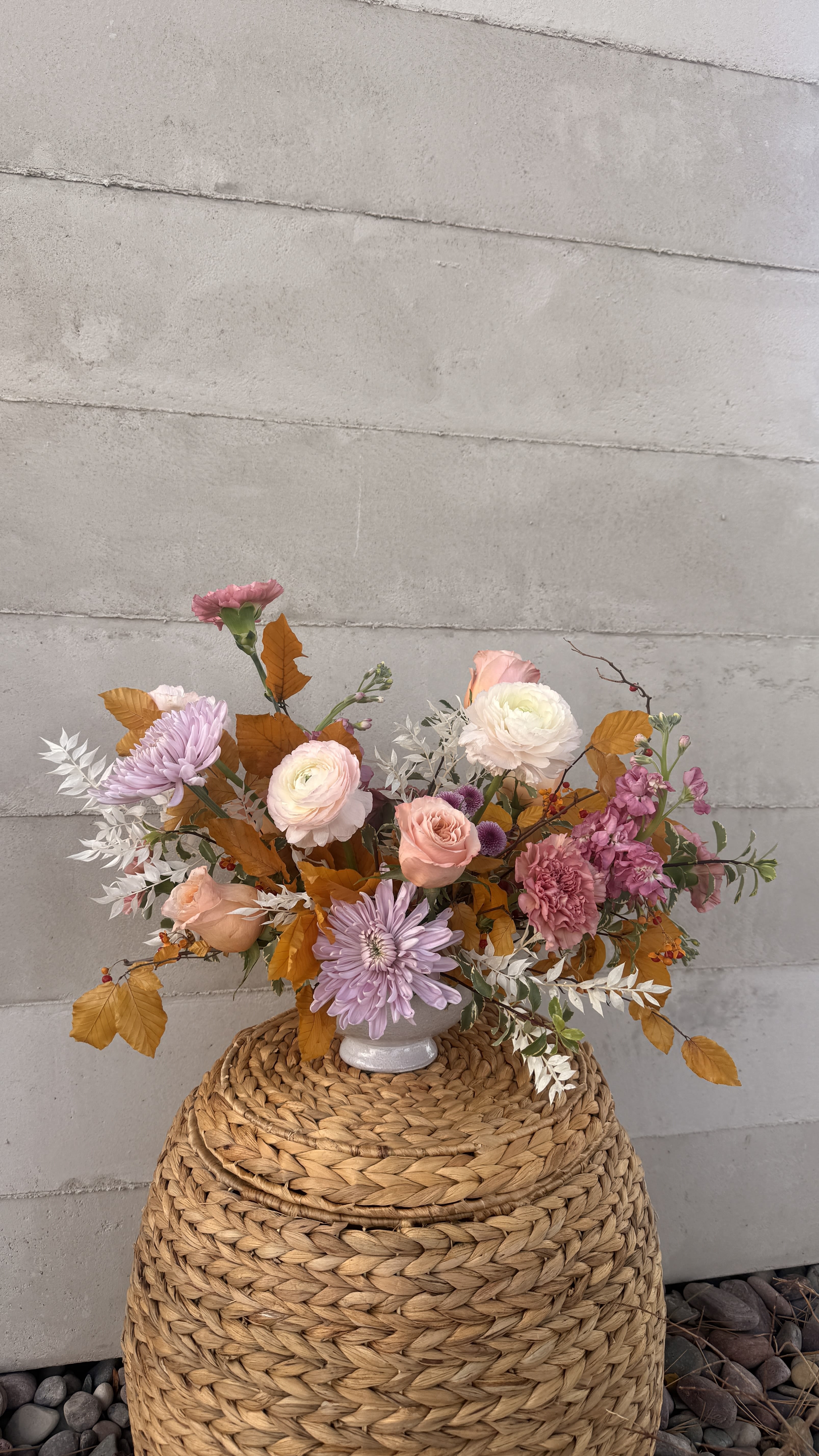 Pastel pink and peach floral arrangement with lavender mums and autumn leaves in a white vase on a woven basket.
