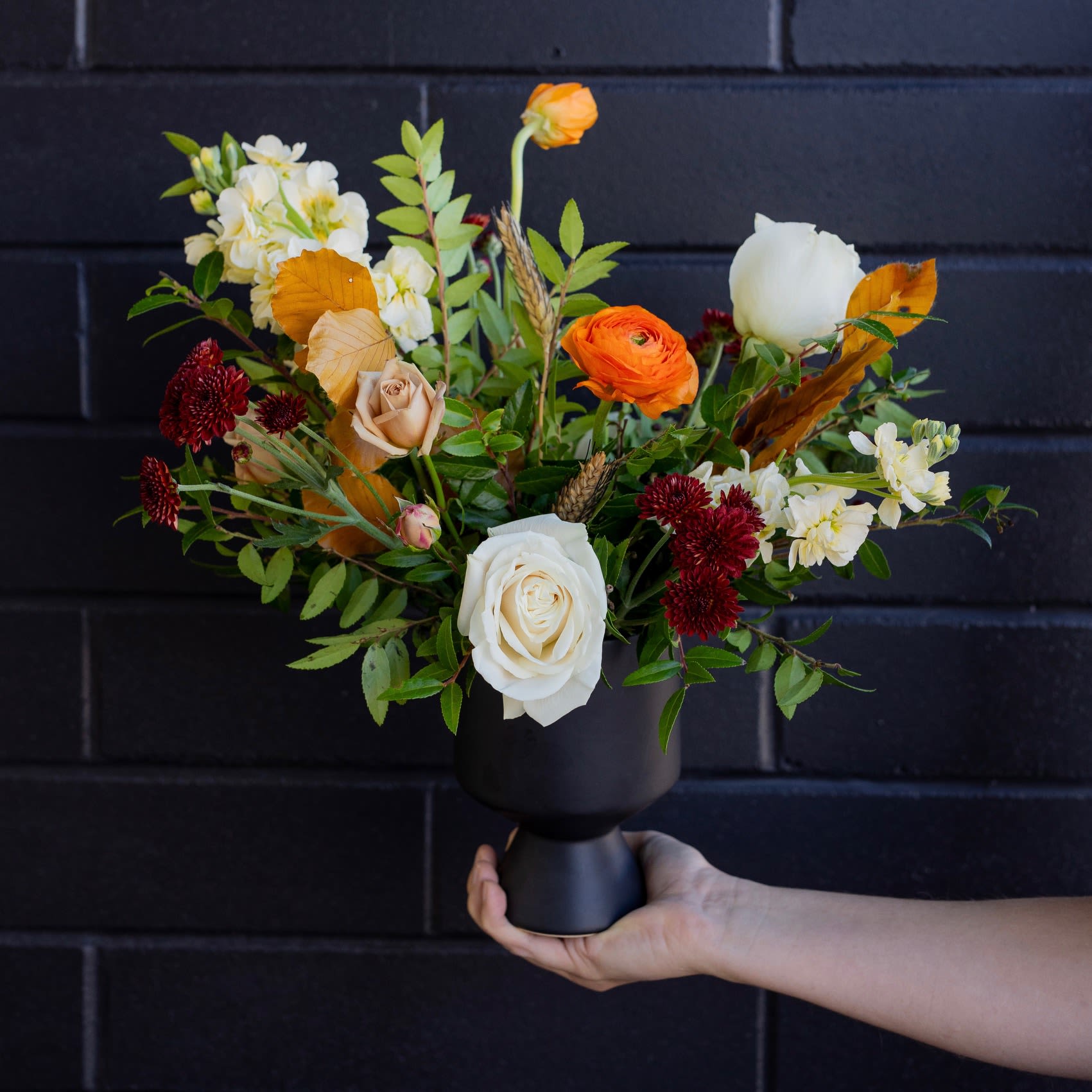Autumn-toned arrangement with white roses, orange blooms, and burgundy flowers in a black pedestal vase.