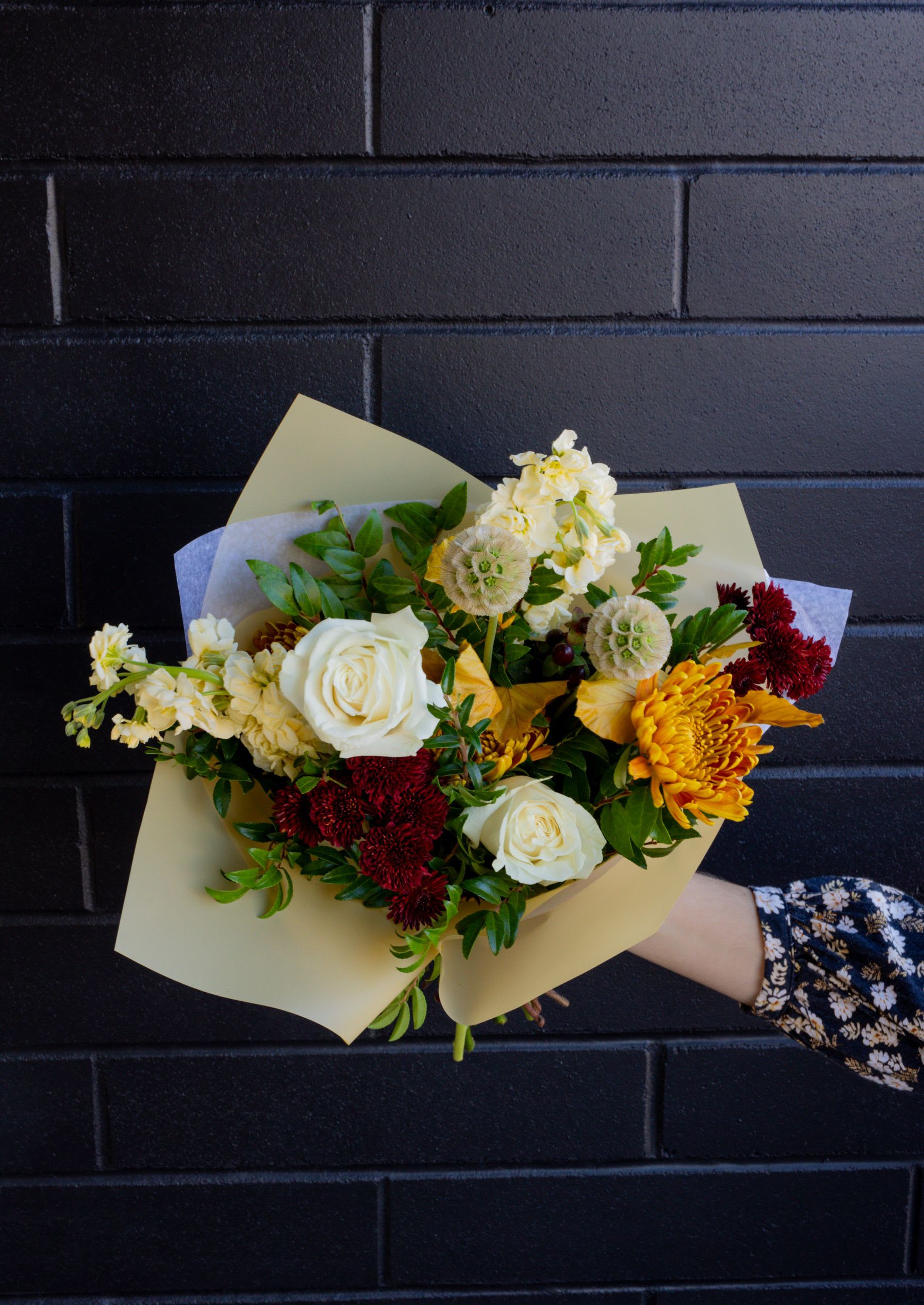 Hand-tied bouquet of white roses, yellow chrysanthemums, and dark red blooms wrapped in pale paper.