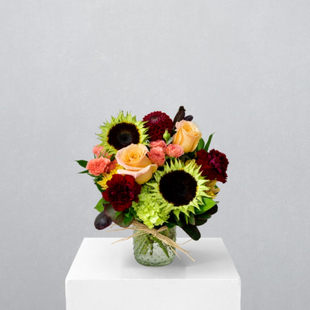 Bouquet of sunflowers, peach roses, and burgundy blooms in a glass vase