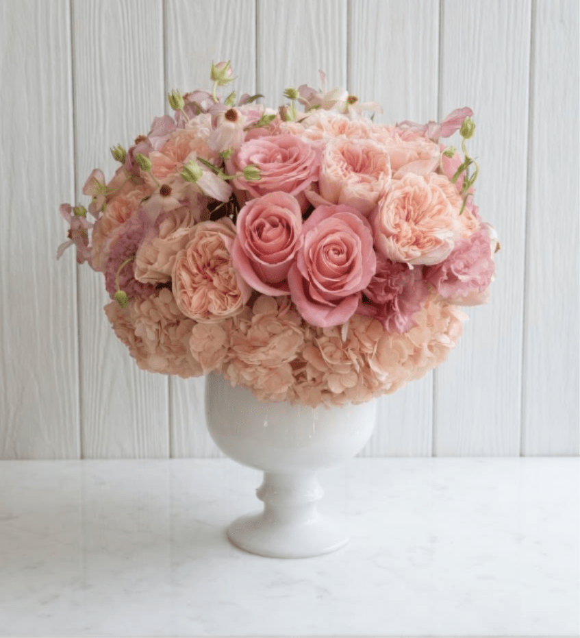 Love Story   - A lush, garden-style arrangement in soft blush and peach tones, featuring velvety roses, ruffled ranunculus, and delicate accent blooms layered over a cloud of hydrangea. Designed in a classic white pedestal vase, it has a full, rounded silhouette that feels both romantic and refined--like a modern take on timeless elegance. 