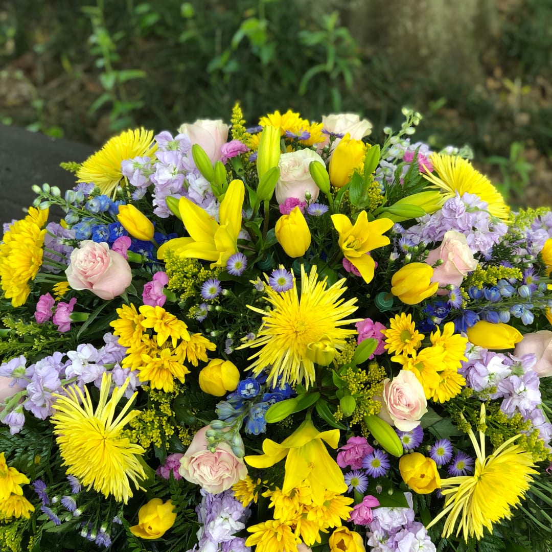 Heavenly Garden Casket Spray - A luminous spray of yellow lilies and chrysanthemums meets soft pink roses in a rounded, abundant design made for sympathy and funeral services. This funeral casket spray brings a gentle, hopeful presence to the room-the kind of arrangement that helps the person you're honoring feel celebrated and remembered with warmth.  Colors of lavender stock, purple daisy-style asters, pale blue delphinium accents, hydrangea touches and mixed carnations are tightly arranged for full visual impact and textured contrast. The palette reads fresh and springlike but remains appropriate year-round; sizes are crafted to drape respectfully across a casket and create a continuous ribbon of blooms from head to toe.  Choose this spray to convey comfort and uplifting remembrance for family, close friends, or a beloved colleague. As a professionally prepared casket piece, it arrives ready to place-no extra assembly required-and should be kept cool and away from direct heat to extend the life of the flowers.
