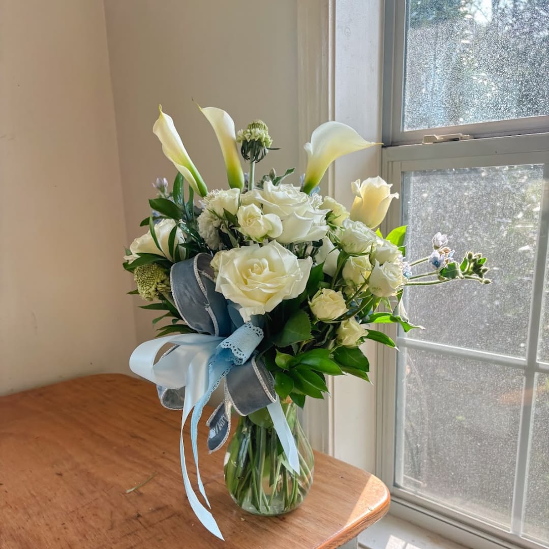 White roses and calla lilies arranged in a glass vase with blue ribbons