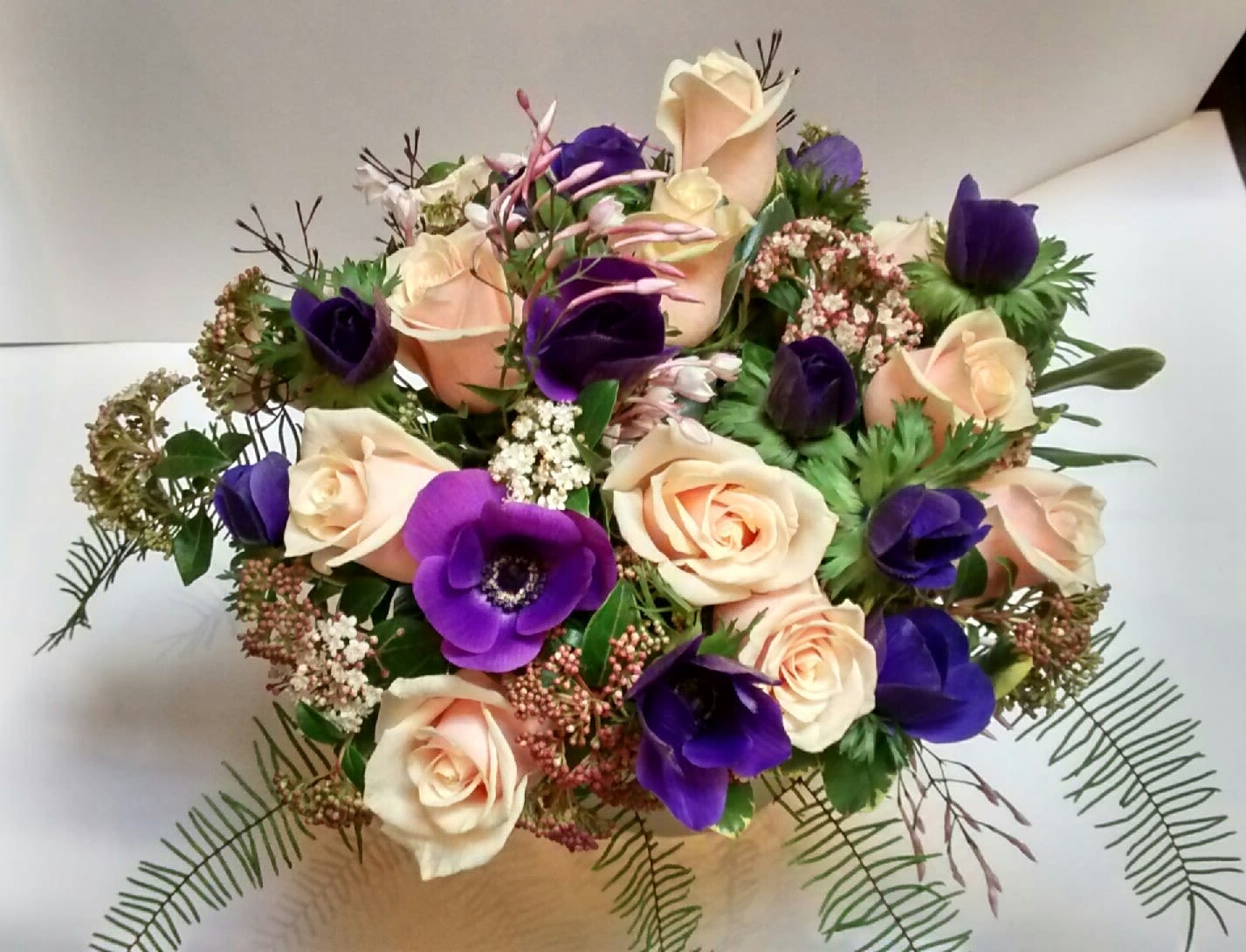 Round arrangement of peach roses and deep purple anemones with airy filler on a white background