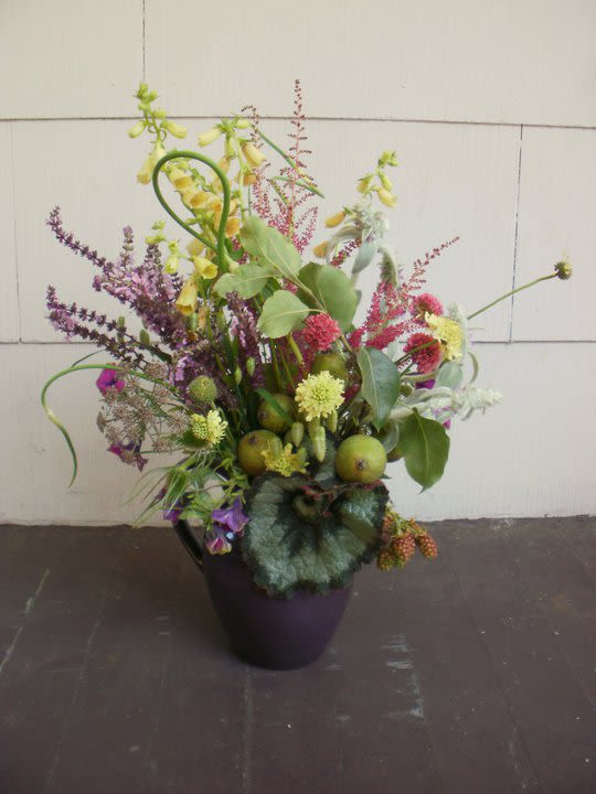 Wild-style arrangement of yellow and purple flowers with green fruits in a dark purple vase
