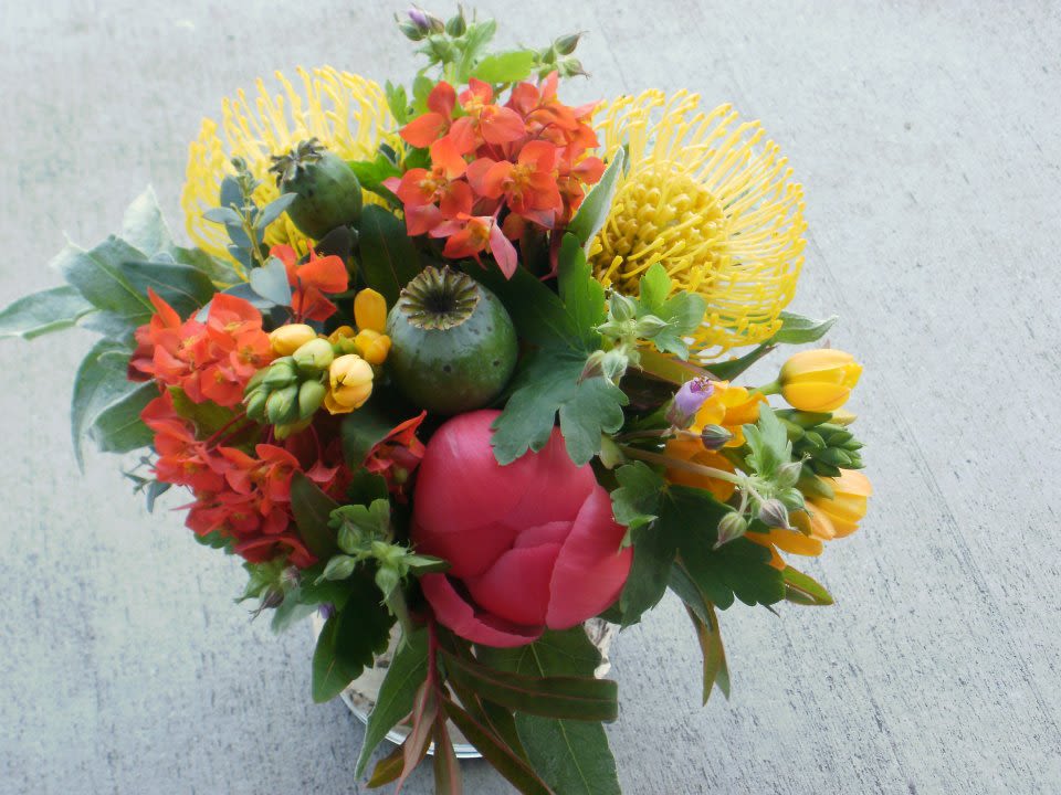Bright mixed bouquet with yellow pincushion protea, orange blooms, and a pink peony in a glass vase