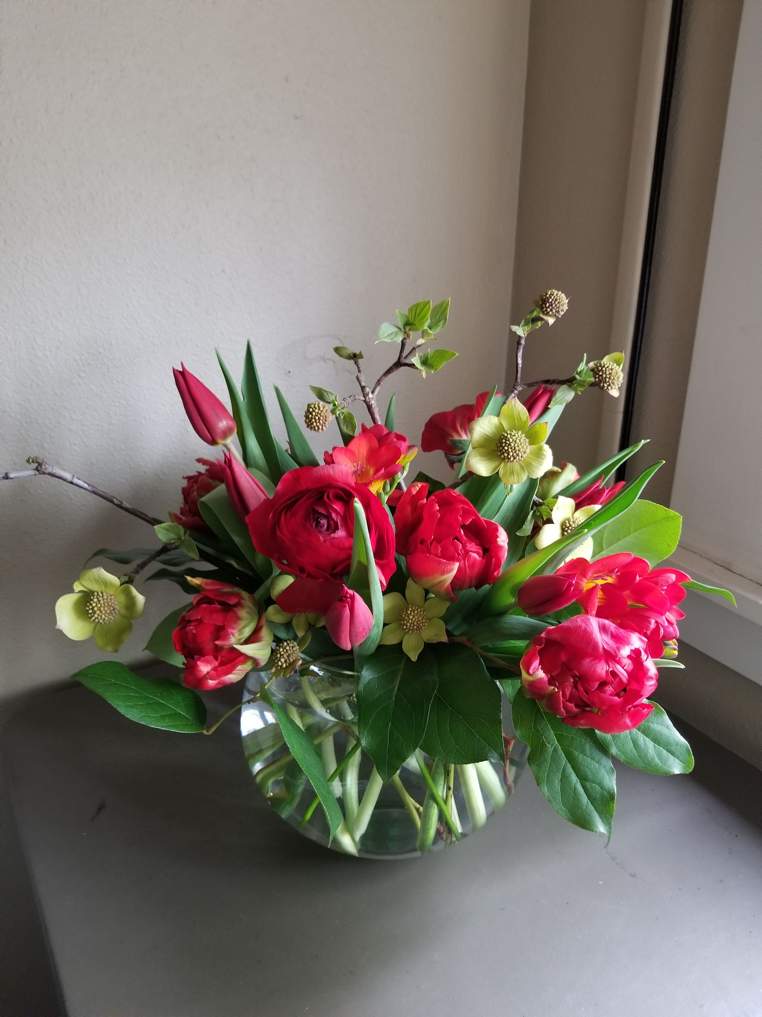 Low round arrangement of red ranunculus and tulips with pale yellow blooms in a clear glass bowl vase.