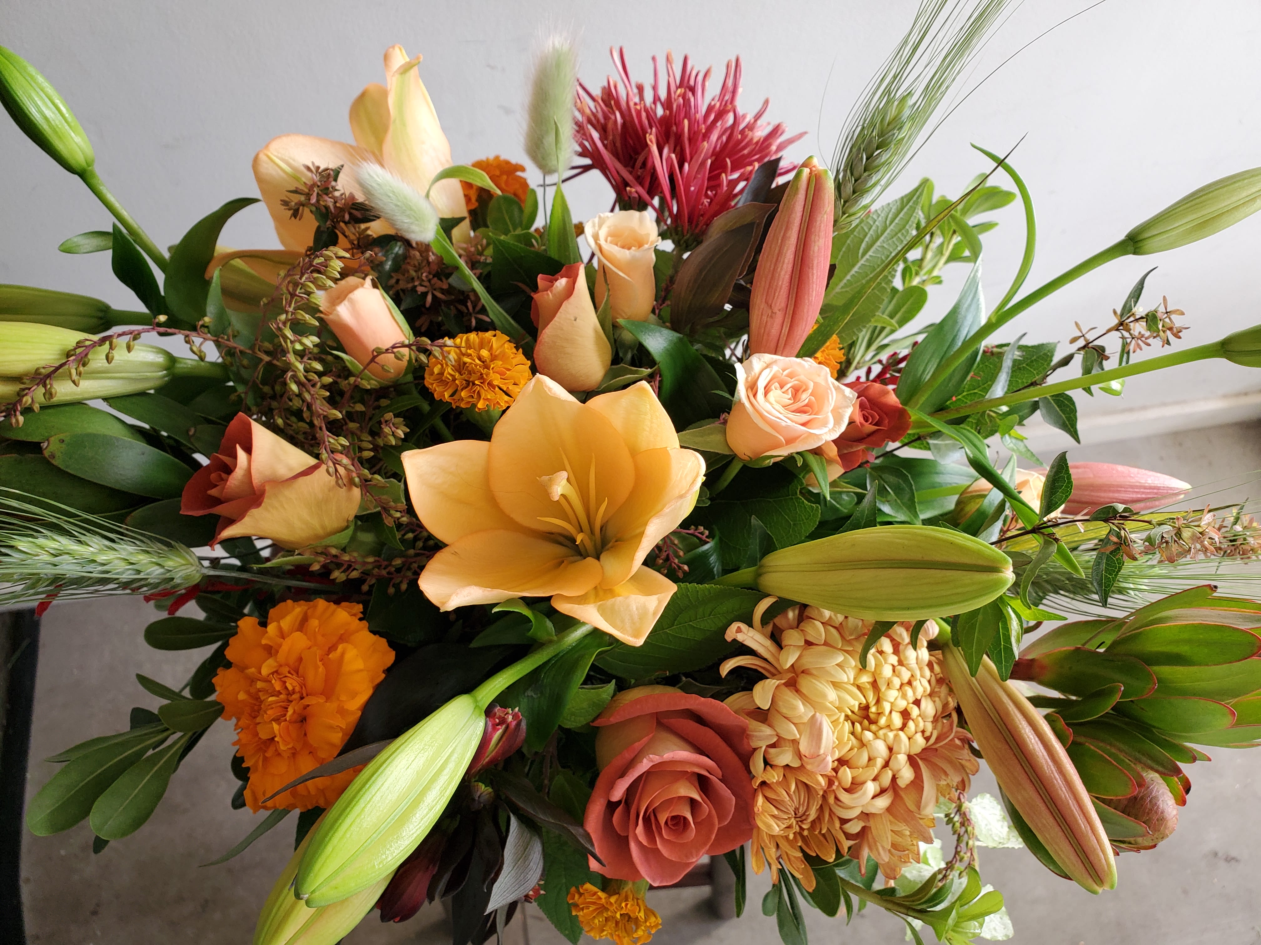 Warm-toned arrangement of peach lilies, roses, marigolds, and mums with buds and textural accents