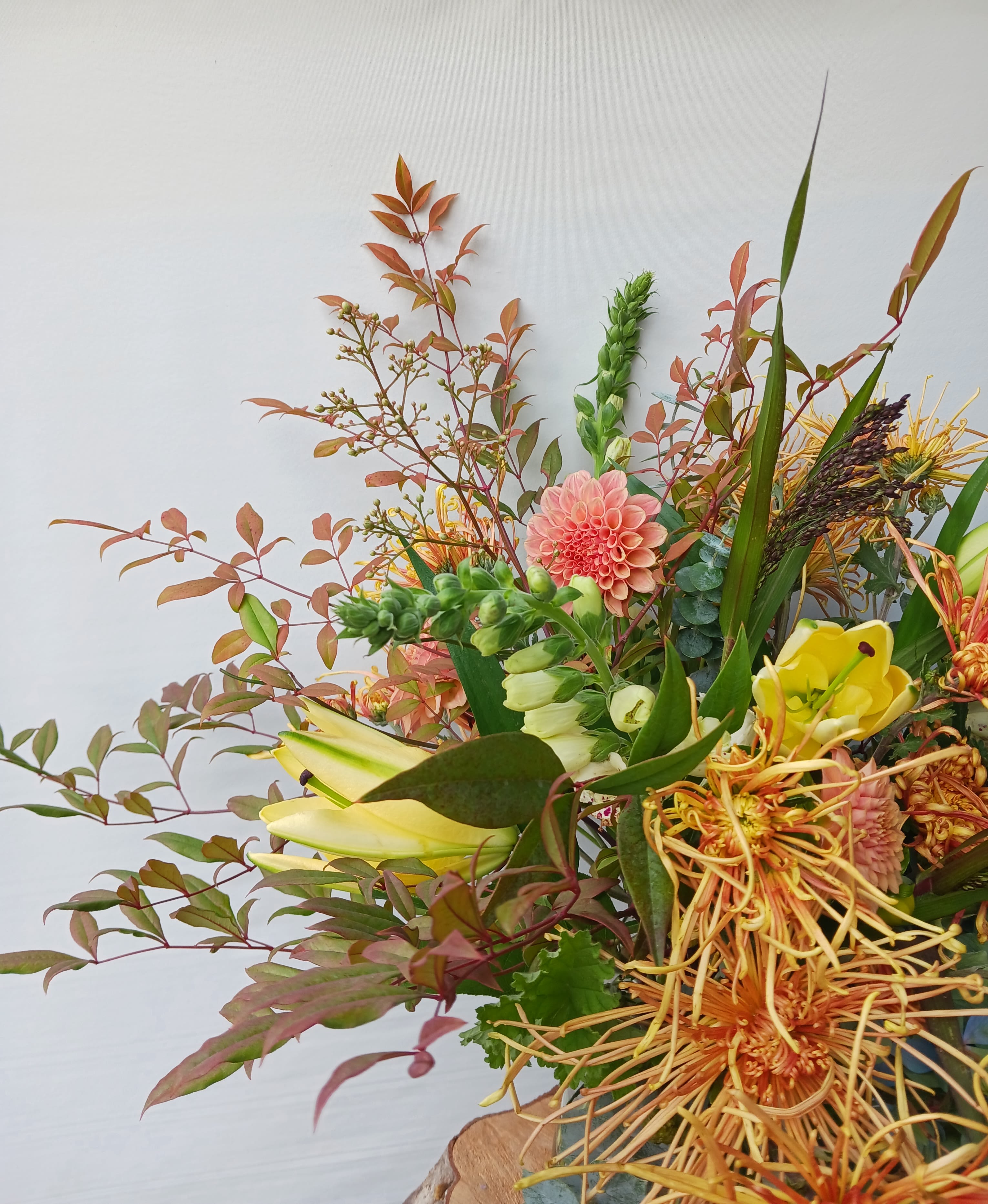 Asymmetrical arrangement with peach dahlias, yellow lilies and orange protea in a vase against a light background