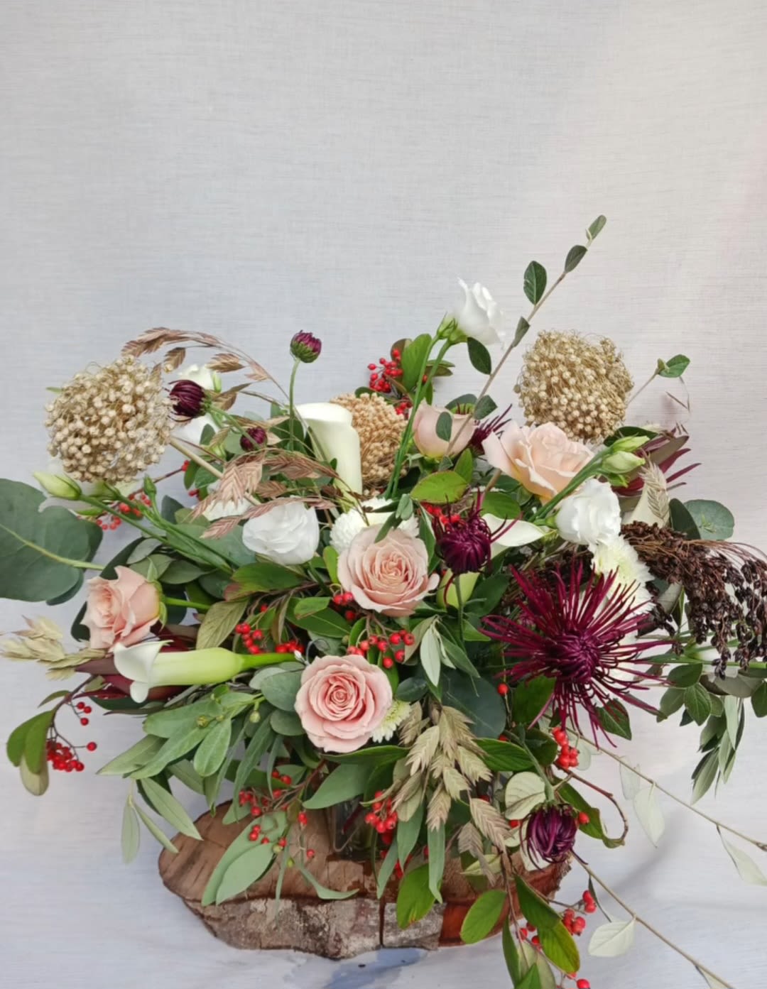 Rustic low arrangement with blush roses, dark red mums, berries, and a candle on a wood base.