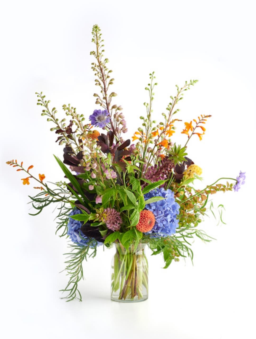 Tall mixed arrangement with blue hydrangeas, orange blooms, and wildflowers in a clear glass vase
