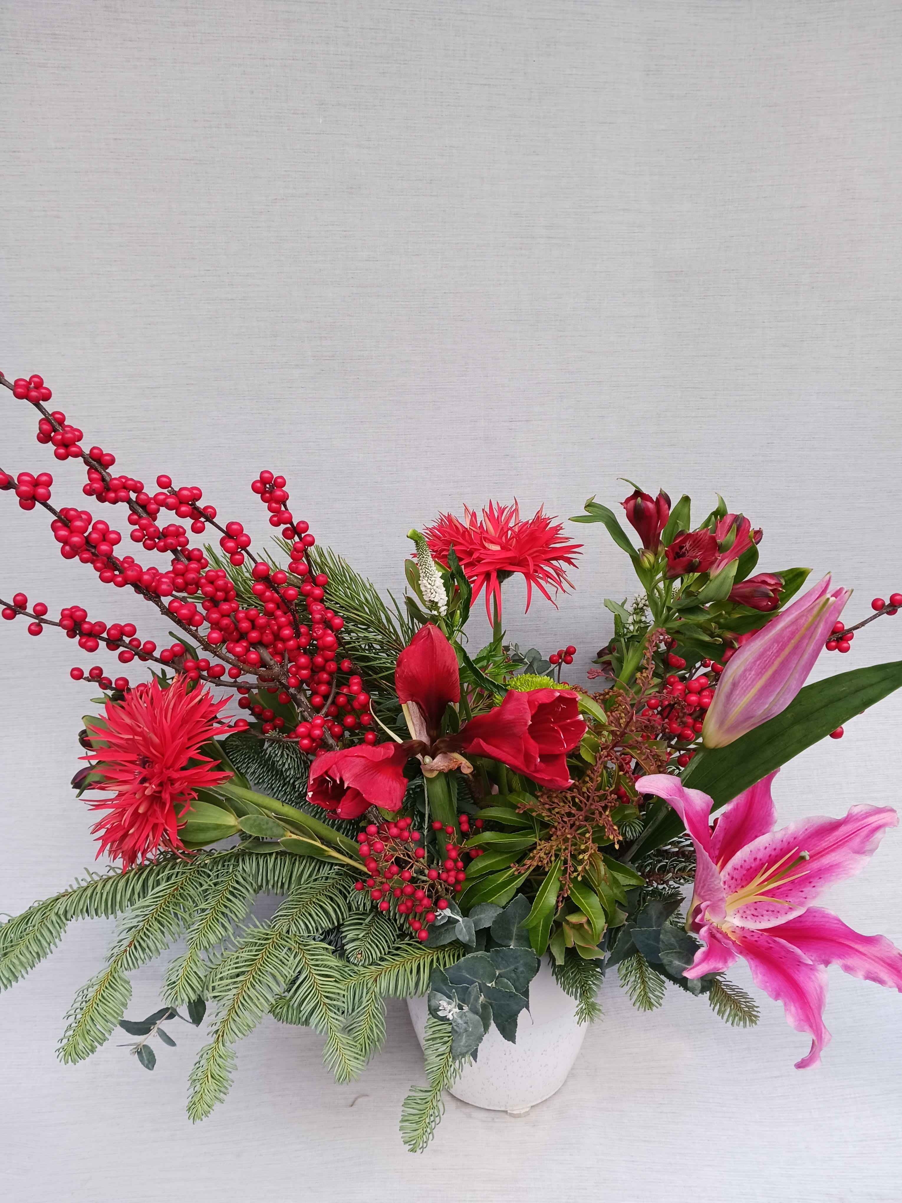 Holiday arrangement with red flowers, berries, a pink lily, and evergreen branches in a white vase