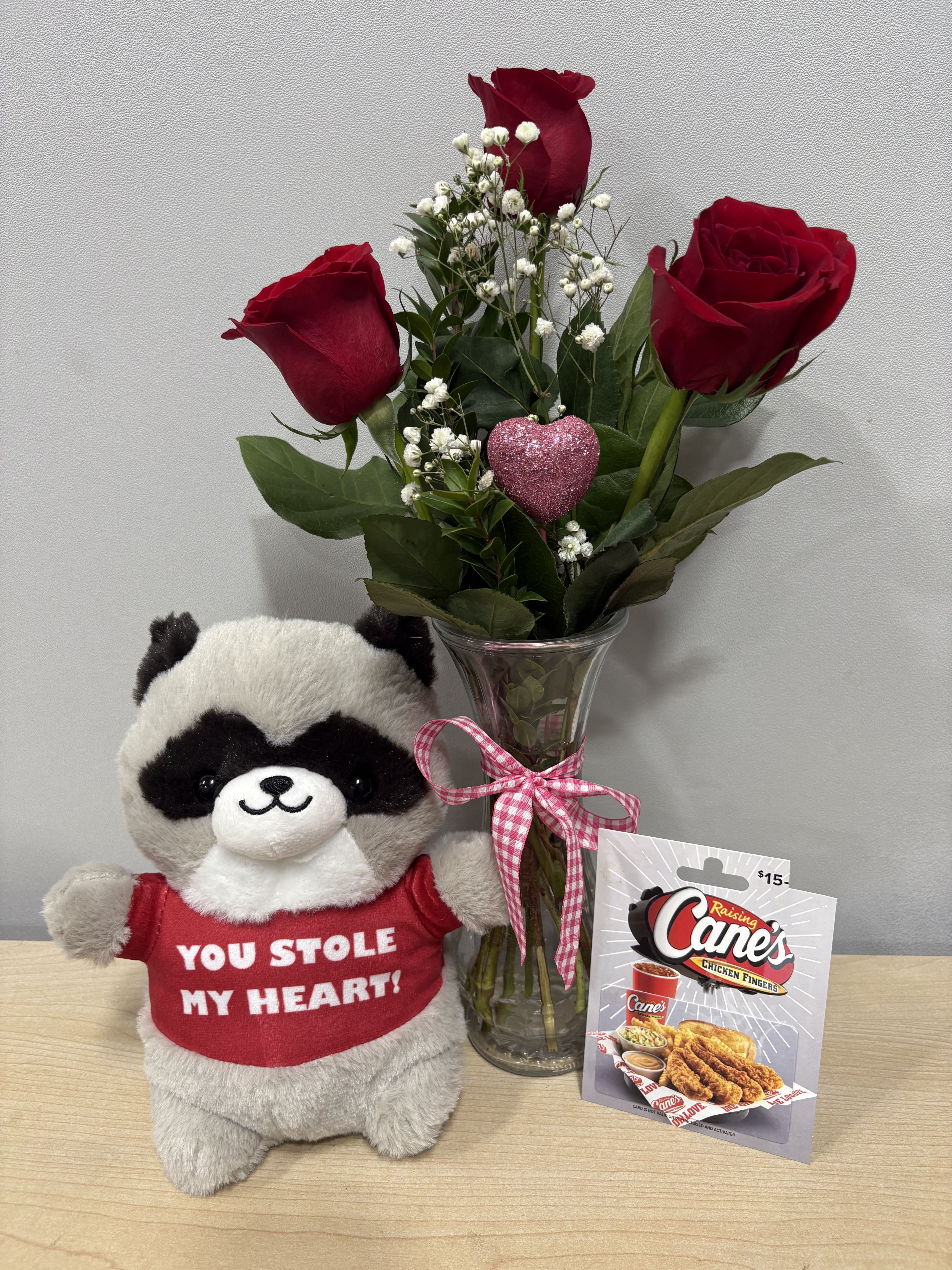 Three red roses in a glass vase beside a plush raccoon and gift card