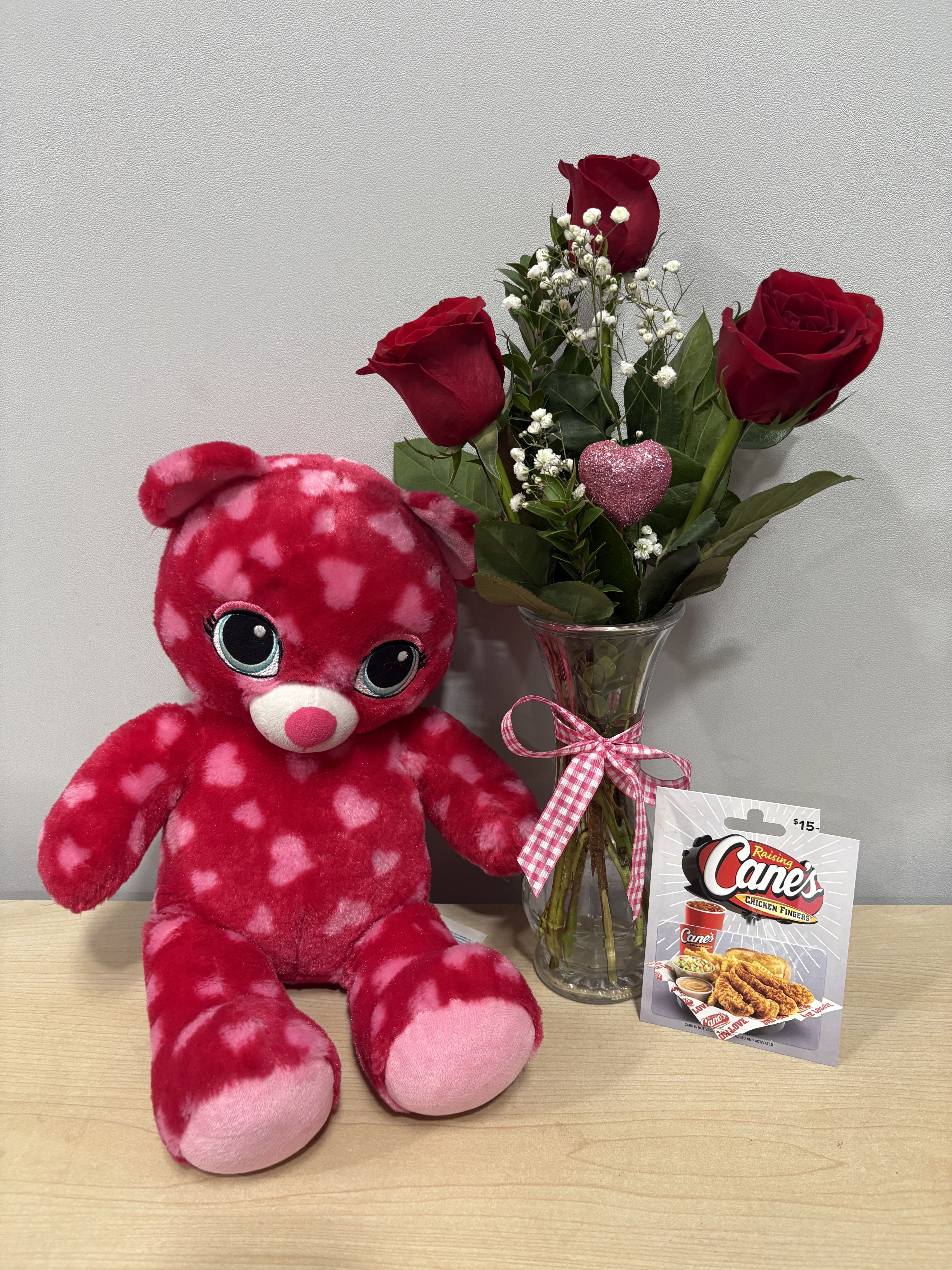 Red roses in a glass vase beside a pink heart-pattern teddy bear and gift card