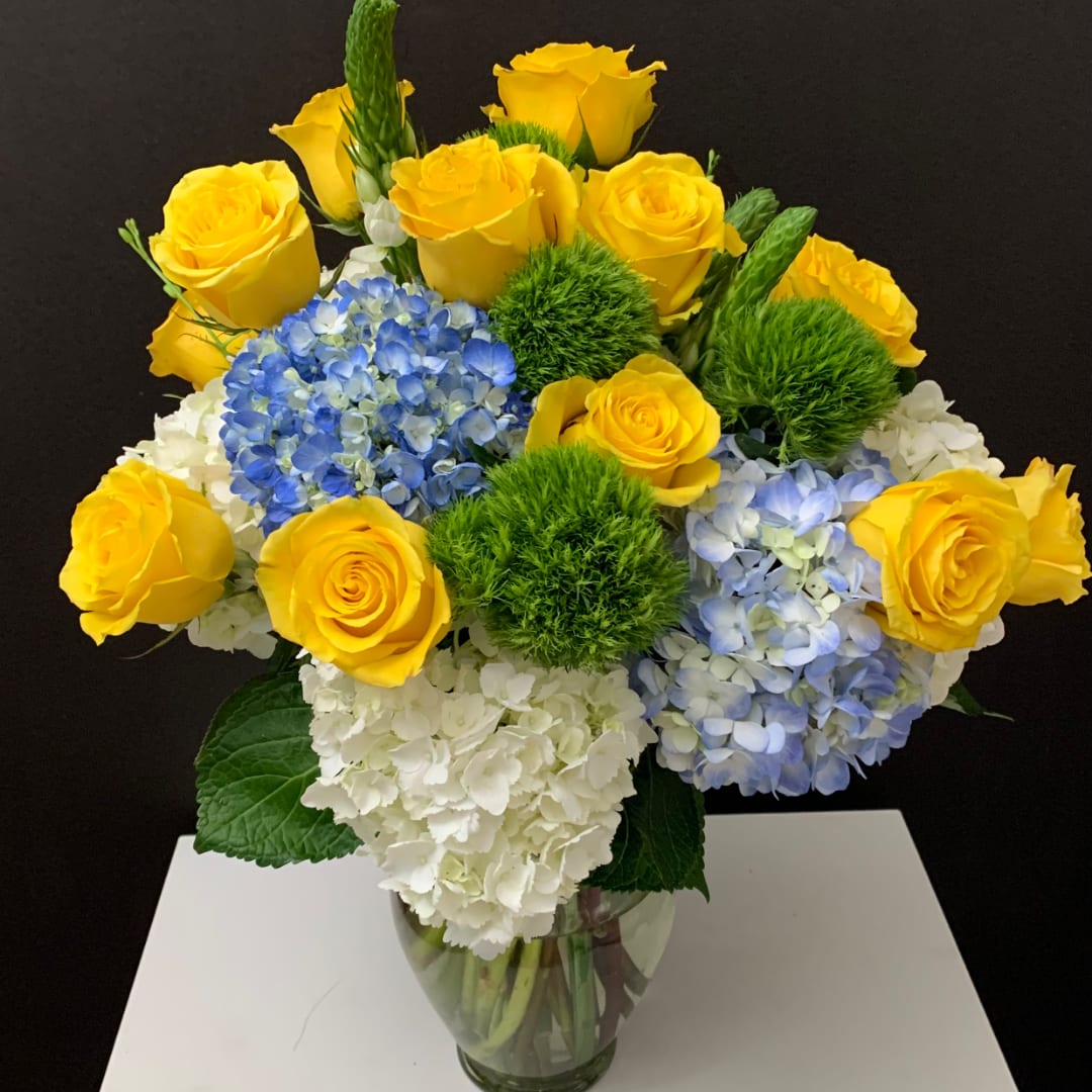 Yellow roses and blue hydrangeas in a glass vase with green pom-pom accents