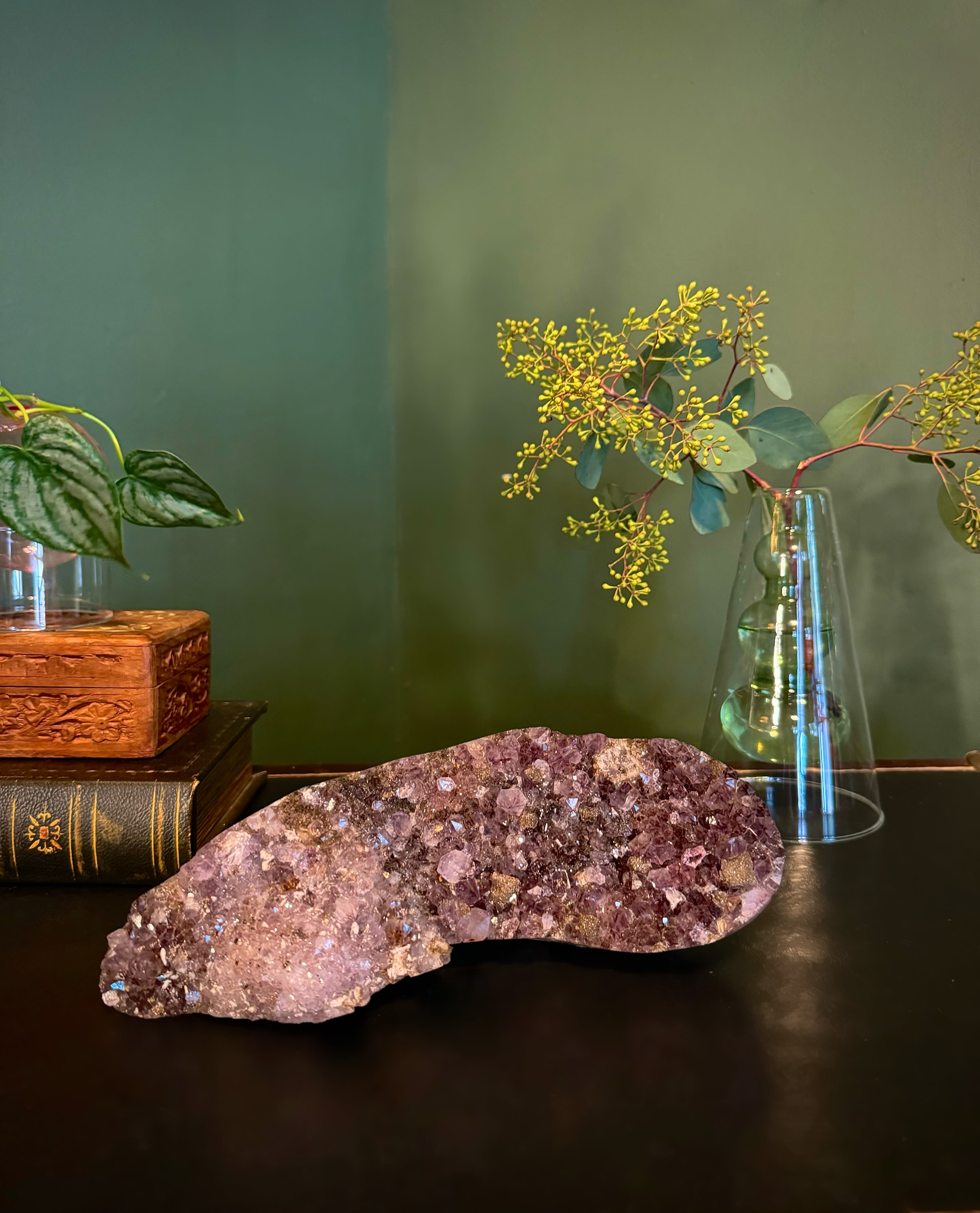 Amethyst crystal cluster on a dark surface with a glass vase and plant stems
