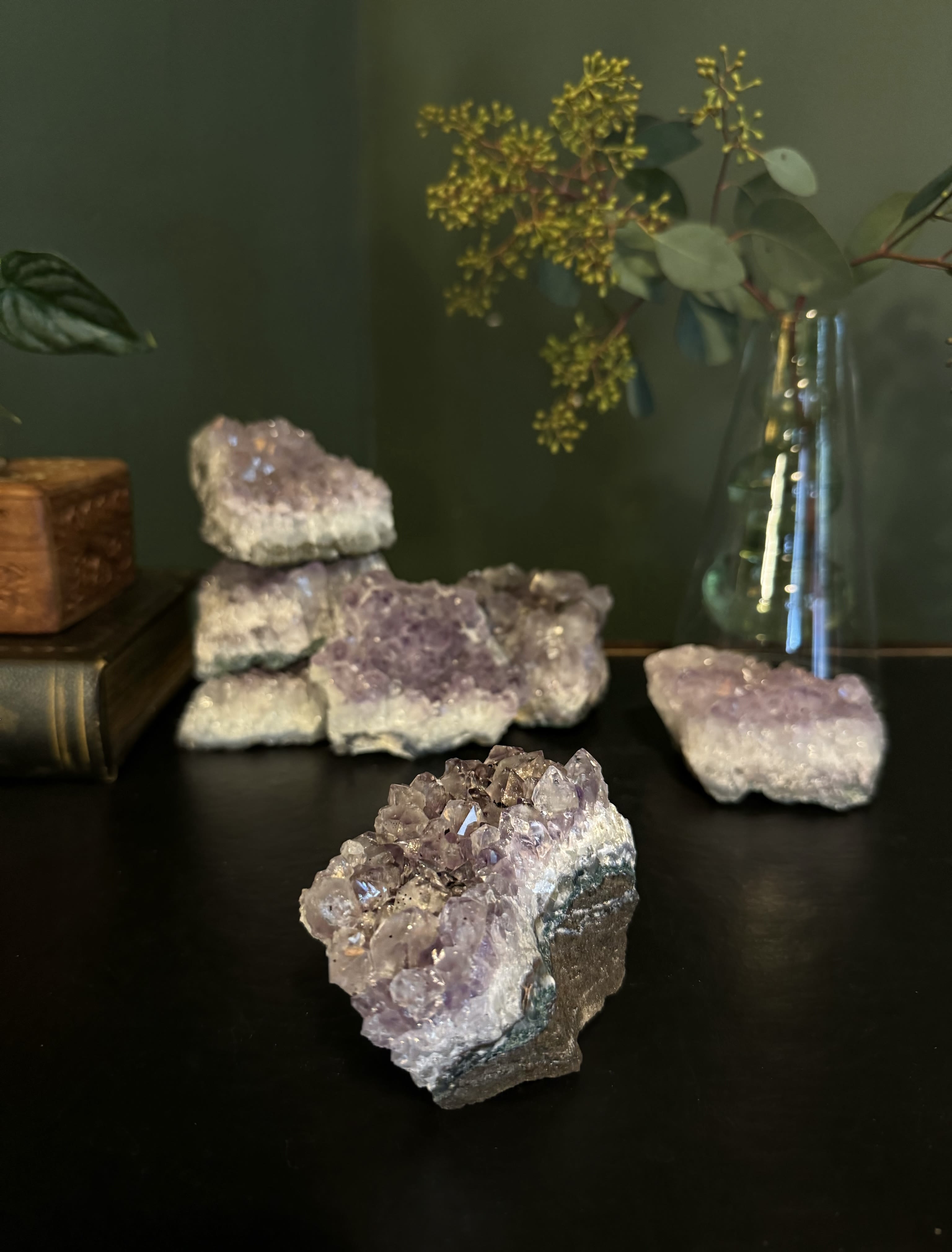 Purple crystal clusters on a dark surface with a glass vase of stems in the background
