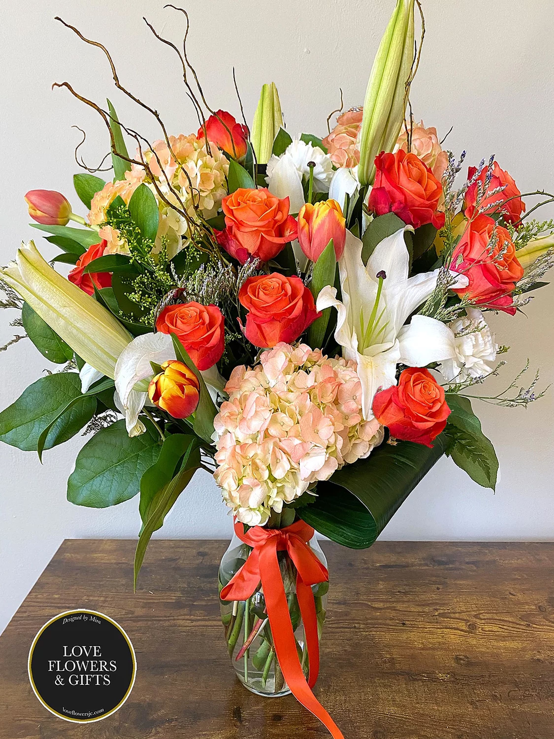 Tall mixed arrangement of orange roses, tulips, white lilies, and peach hydrangeas in a clear vase with orange ribbon