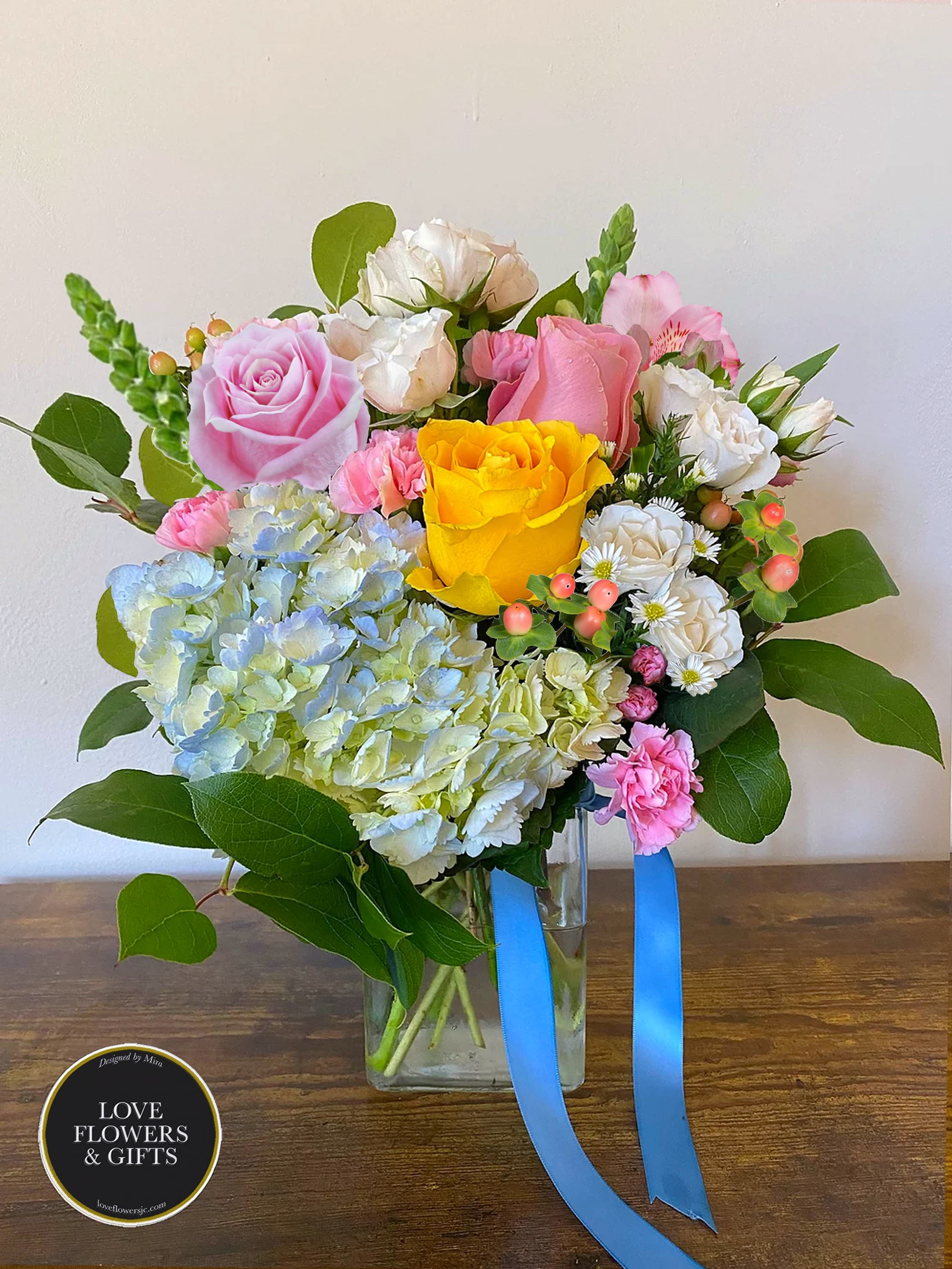 Mixed bouquet of pink and yellow roses, hydrangea, and carnations in a clear glass vase with blue ribbon