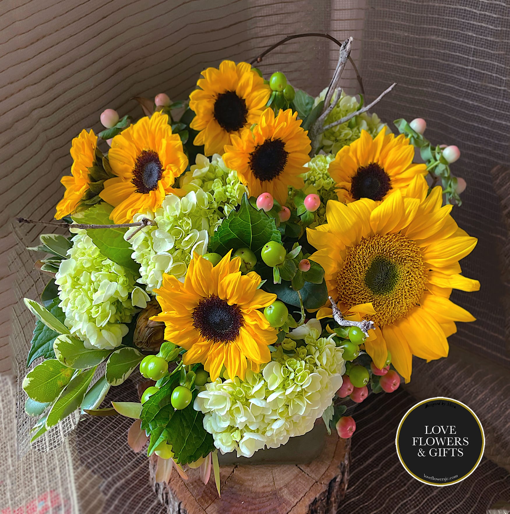 Arrangement of yellow sunflowers and pale green hydrangeas in a low vase on a wood slice