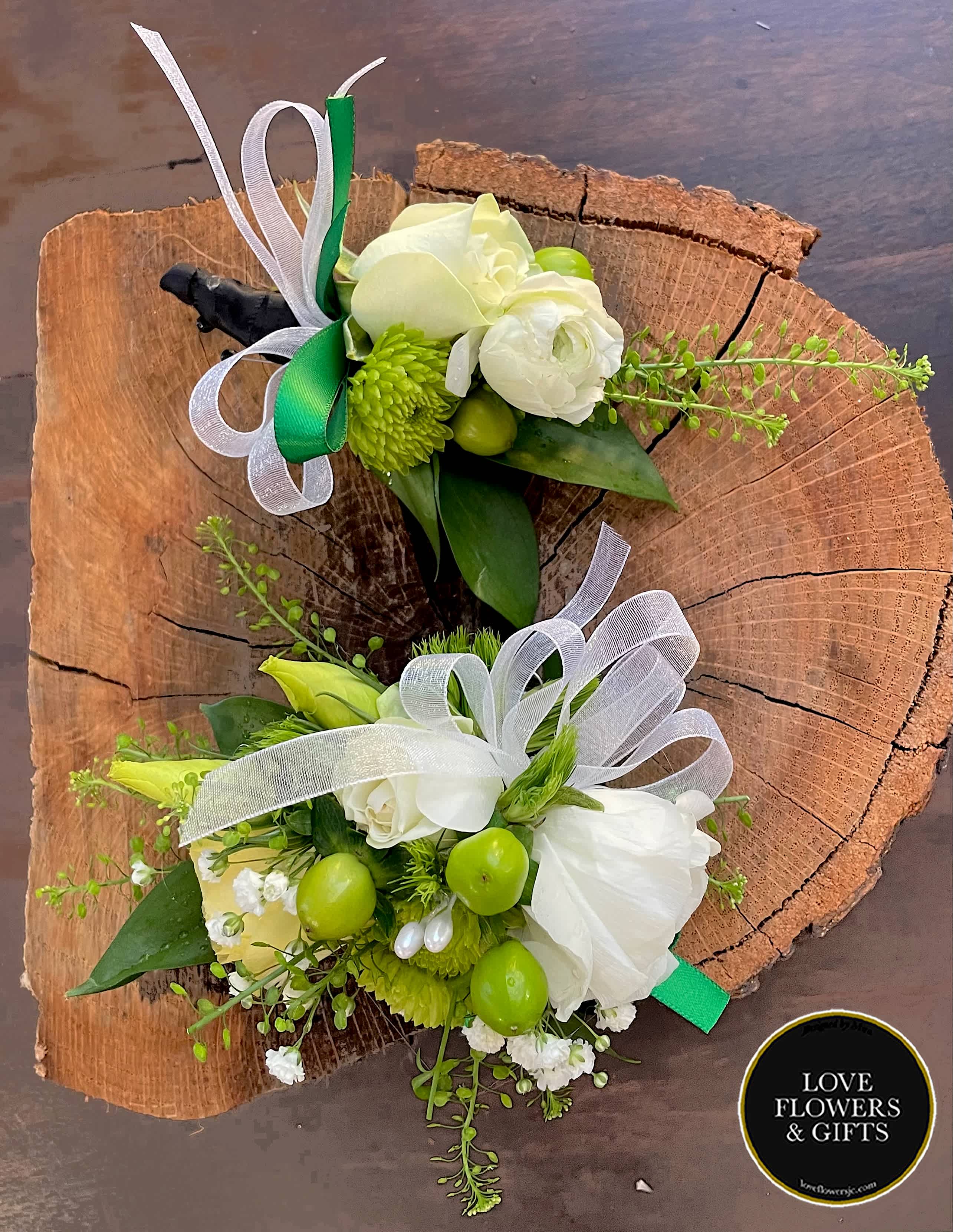 Pair of white and green floral boutonnieres with ribbons displayed on a rustic wood slice