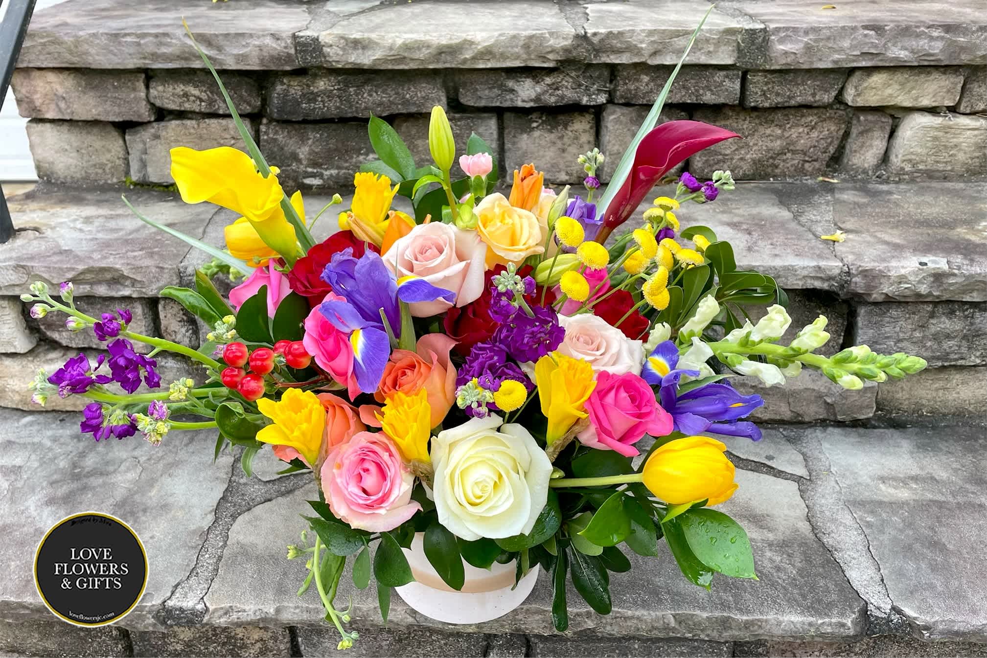 Low round arrangement of colorful roses, tulips, irises, and other blooms in a white container.
