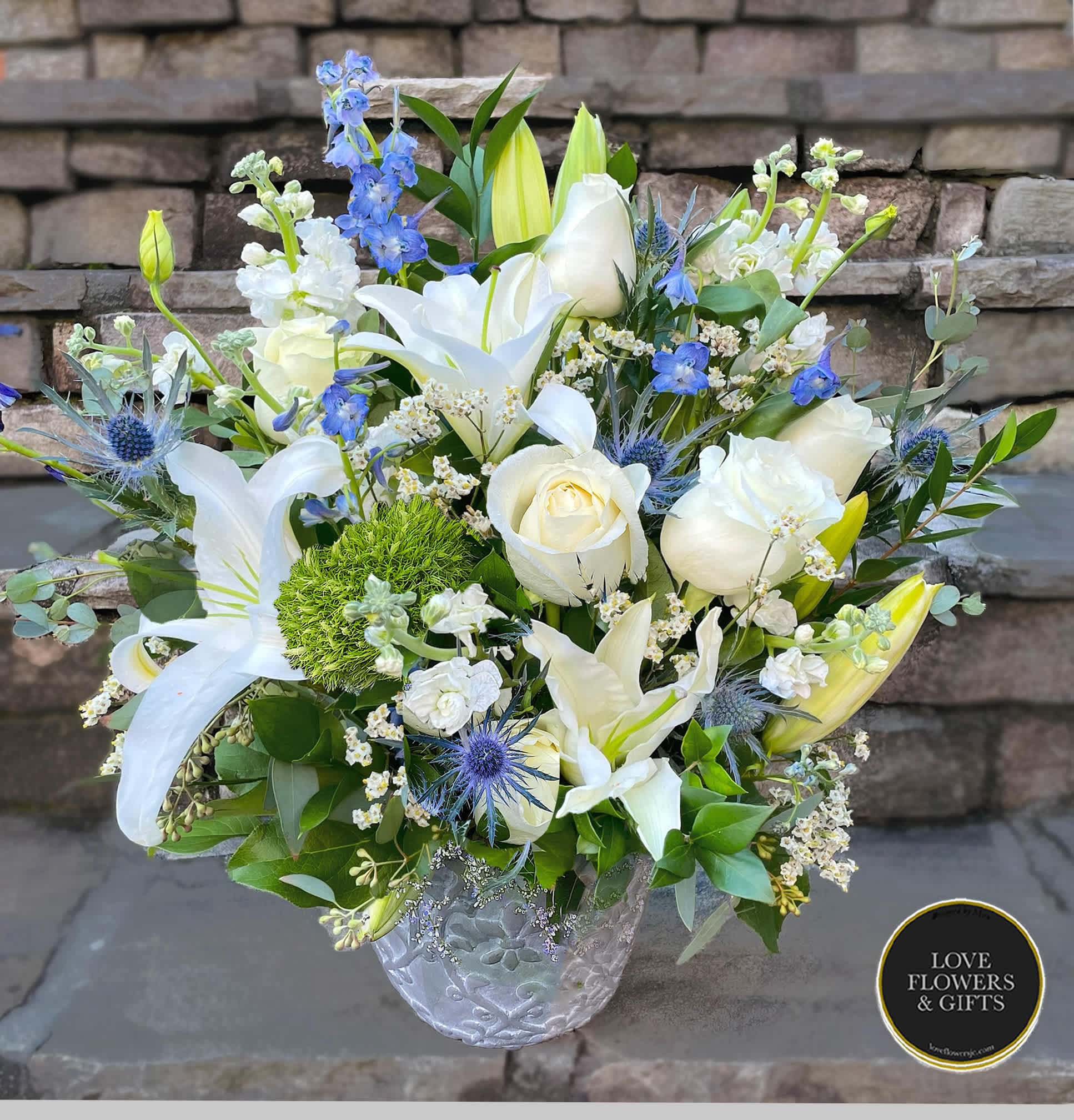 White lilies and roses with blue accents arranged in a clear patterned glass vase
