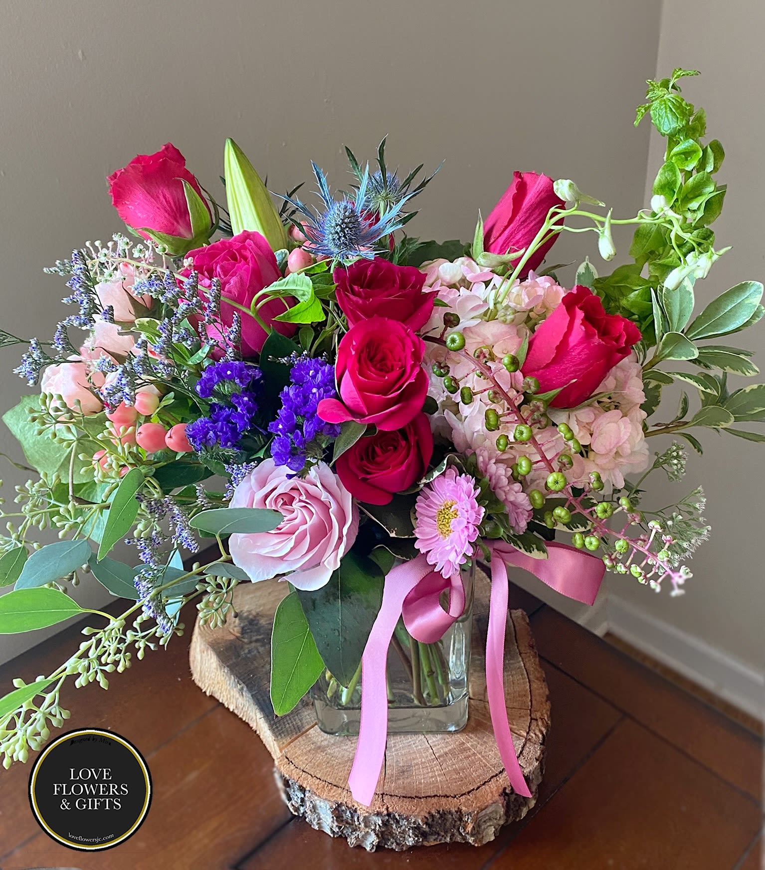 Pink and purple mixed flower arrangement with roses and hydrangea in a glass vase on a wood slice stand.