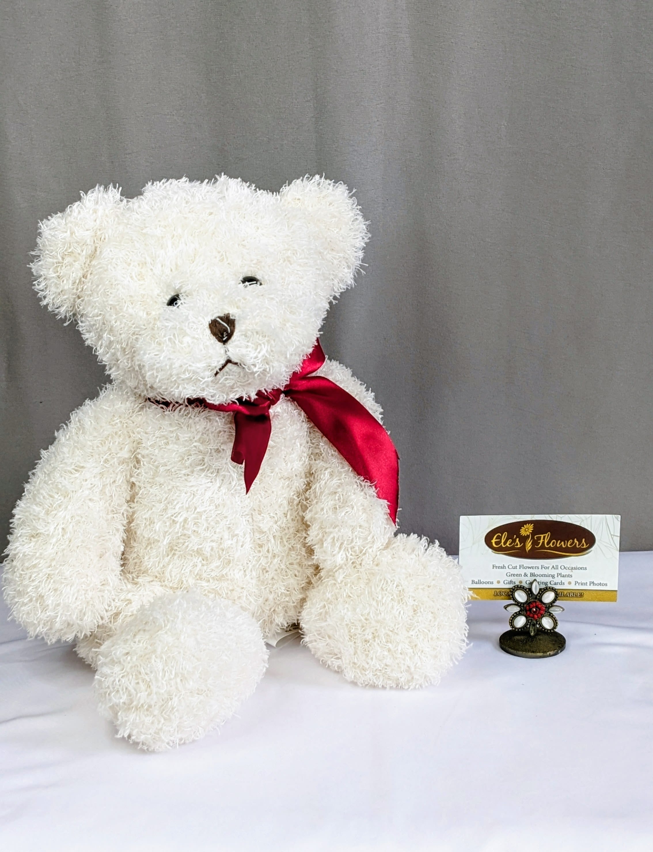 White teddy bear with a red ribbon sitting beside a florist card
