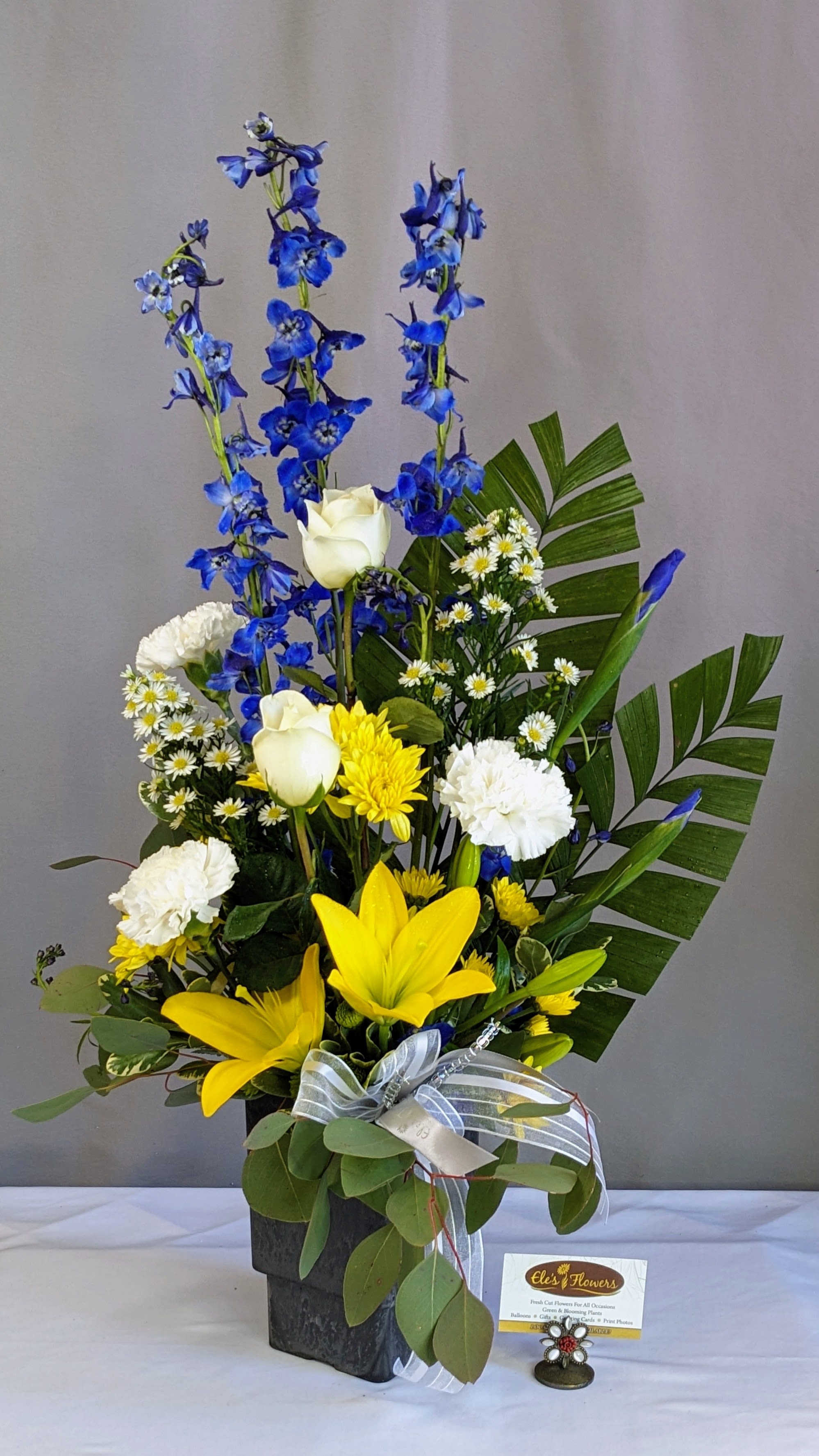 Contemporary Blues, Whites and Yellow - A lovely assortment of blues, yellows and white. Towering blues cascade into whites and yellows. Our design has a contemporary feel and design. Perfect for celebrations of life and funeral services.   The designs we create are custom and the blooms and containers will vary. We use the freshest product available to create the perfect floral combination for the recipient! If you have any special requests, please add those in the "special instructions" portion of your order to help us dial in your vision. Otherwise, our skilled designers, Brett &amp; Sara, will take it from here! Please note that the photos are examples only – floral selection will vary but colors will remain the same.   Approx. Dimensions: 20”+H x 16”+W