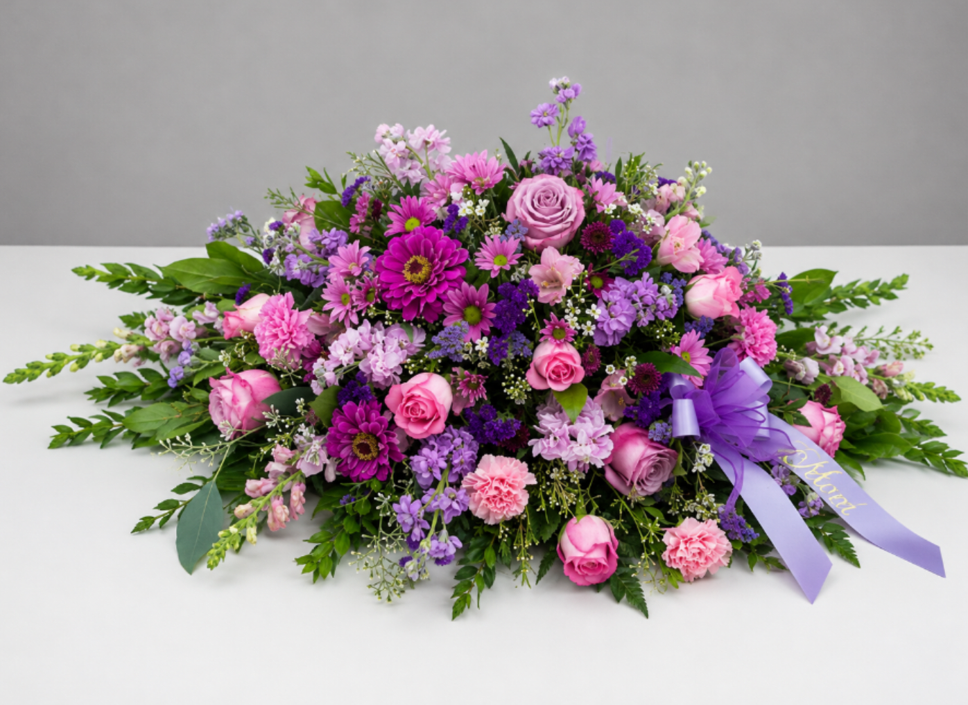 Compassionate Lavender Blooms Casket Spray - A graceful tribute designed to express love, comfort, and heartfelt remembrance. Compassionate Lavender features a lush gathering of soft pink and lavender blooms accented with rich purple tones, delicate textures, and abundant greenery. A coordinating lavender remembrance ribbon adds a meaningful finishing touch to this elegant casket spray.  Thoughtfully designed to convey peace and compassion, this arrangement offers a beautiful way to honor a cherished life while surrounding family and friends with the gentle beauty of flowers.  Flower varieties may vary based on seasonal availability while maintaining the soft pink, lavender, and purple color palette and overall style of the arrangement.