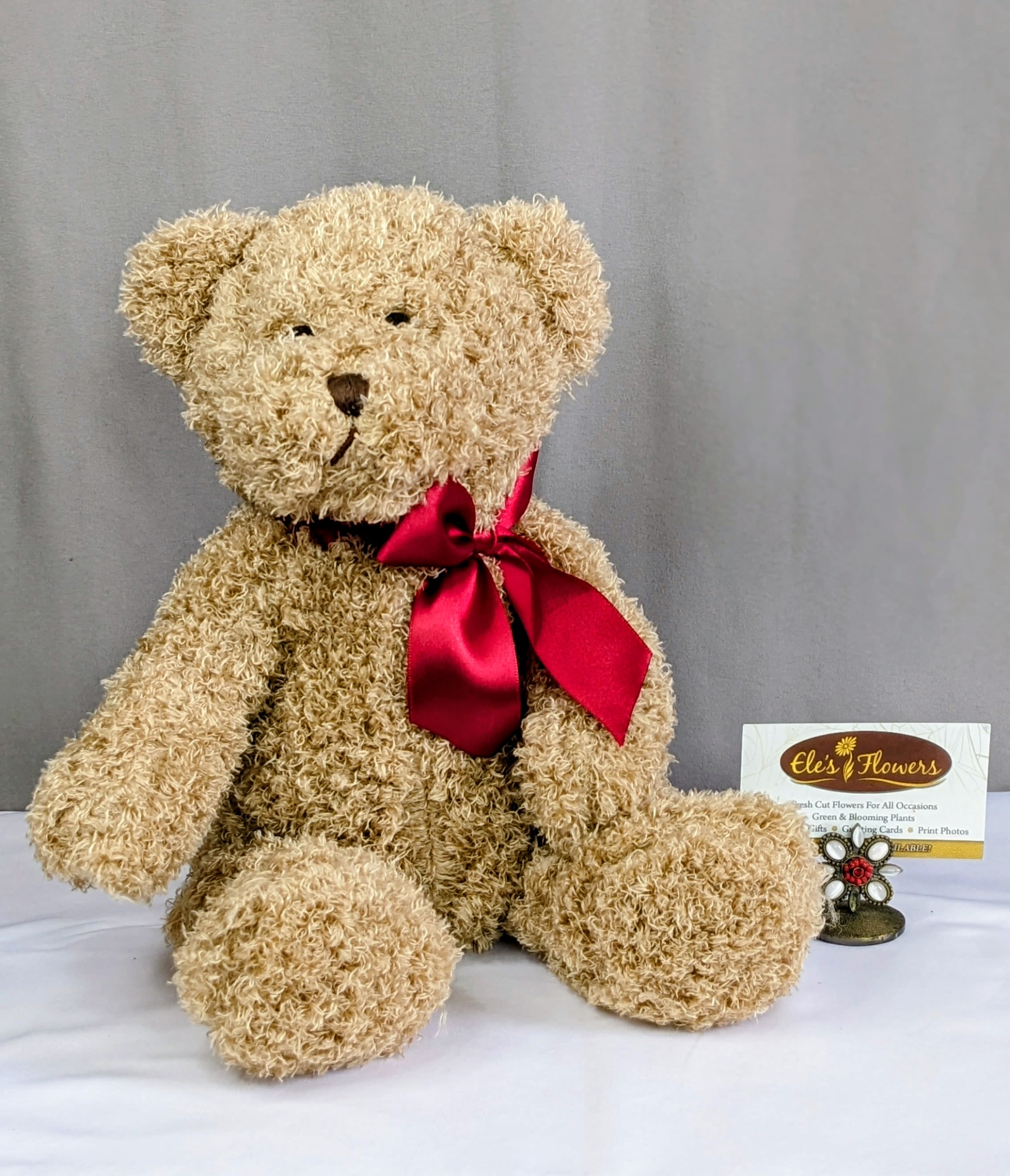 Tan plush teddy bear with a red satin bow sitting beside a florist card