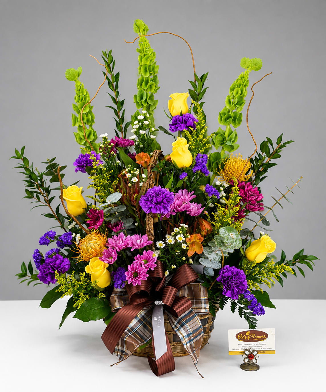 Basket arrangement with yellow roses, purple and pink blooms, and tall green accents
