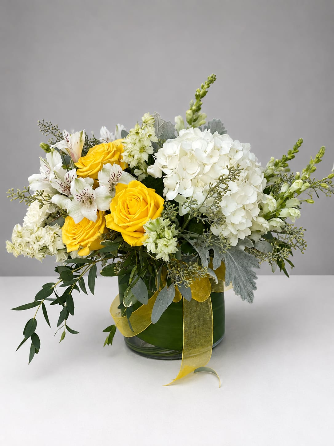 Yellow roses and white hydrangeas in a glass vase with a yellow ribbon
