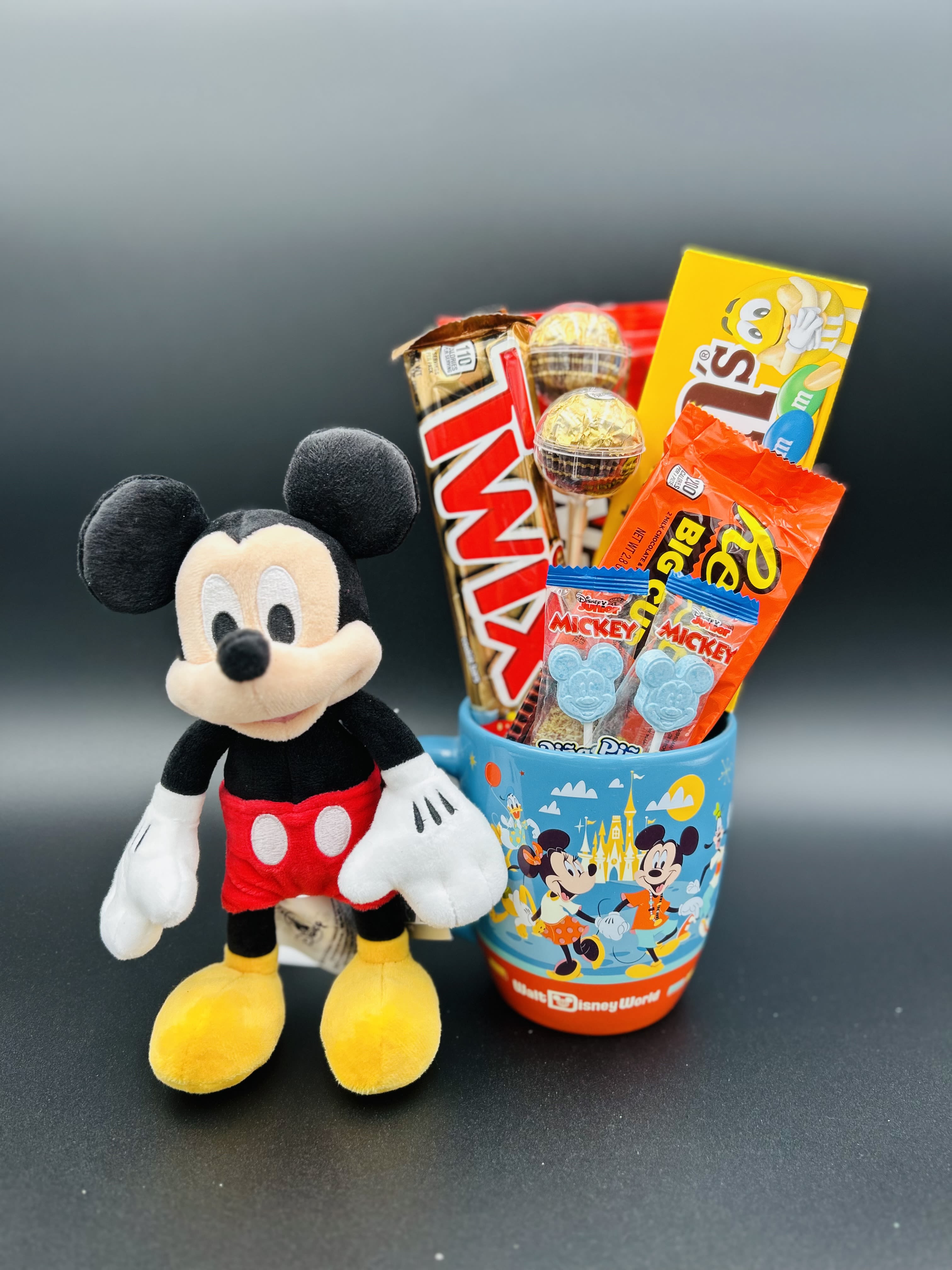 Mickey Mouse plush beside a Disney-themed cup filled with candy bars and lollipops