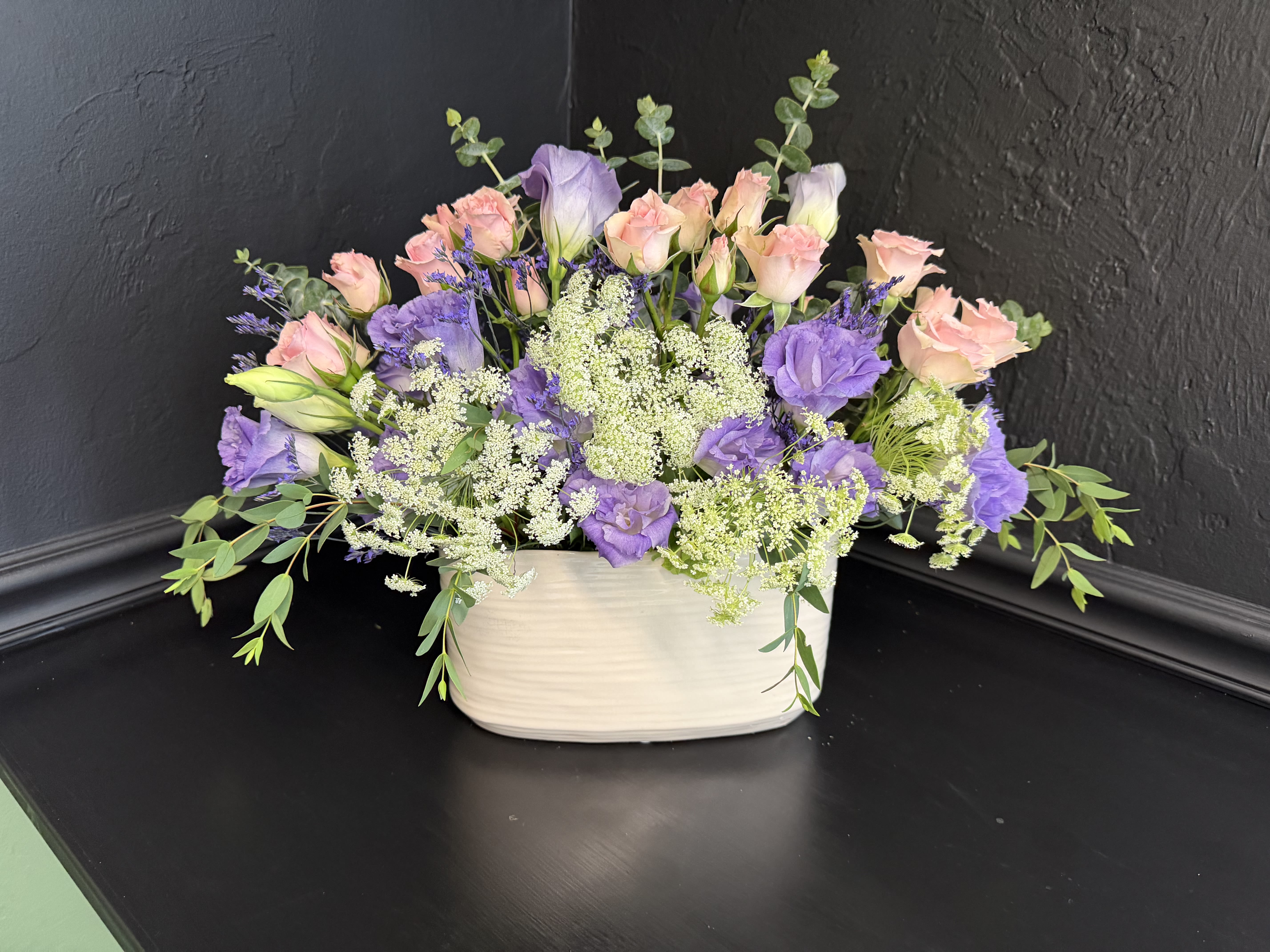Gentle Love - Soft lavender and blush pink blooms mingle in a low, wide composition that reads like a gentle garden tucked into a white ceramic vase-perfect as a birthday bouquet, anniversary arrangement, or a thoughtful "thinking of you" gift. Lavender Whisper uses pastel purples and pinks to create a romantic, calming impression so the person you're sending it to feels quietly celebrated and seen.  This floral arrangement pairs blush pink roses with lilac lisianthus and other lavender-toned blooms, accented by airy white filler flowers and trailing greenery for a loose, garden-style silhouette. The flowers are arranged in a white rectangular ceramic vase that keeps the design compact and tabletop-friendly while letting stems and texture show; colors are soft purples, pinks, whites, and fresh green foliage.  Ideal for dining centers, or a desk to brighten someone's day, Lavender Whisper conveys warmth without fuss-an elegant everyday floral gift for birthdays, anniversaries, thank-yous, or just because. To care for: keep out of direct heat for longer enjoyment.