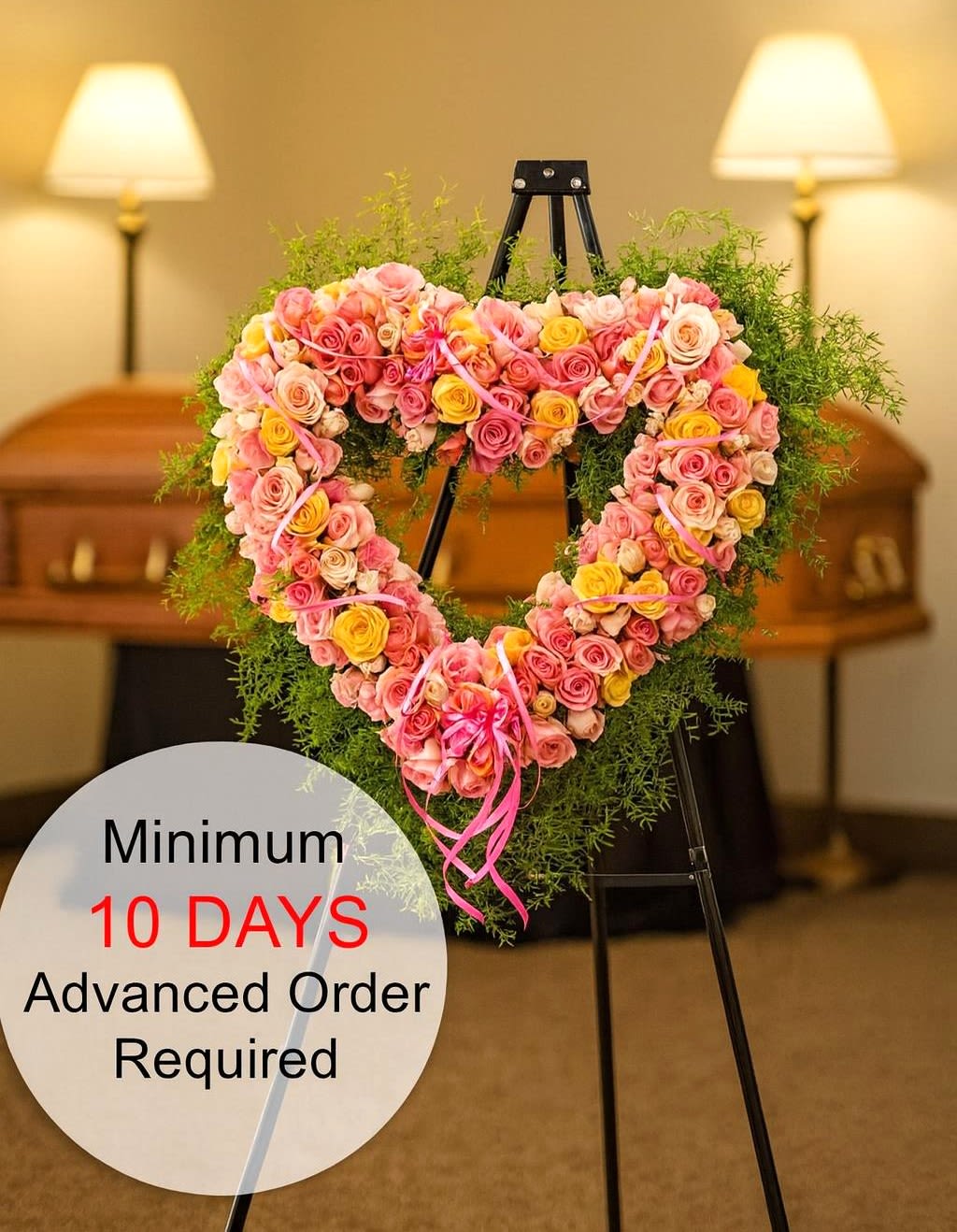 Heart-shaped wreath of pink and yellow roses on a metal easel in a funeral setting.
