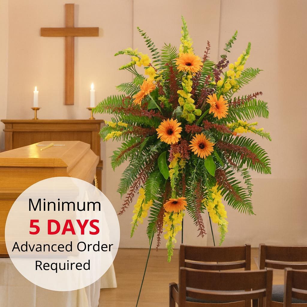 Tall sympathy easel spray with orange gerbera daisies, yellow blooms, and ferns beside a casket.