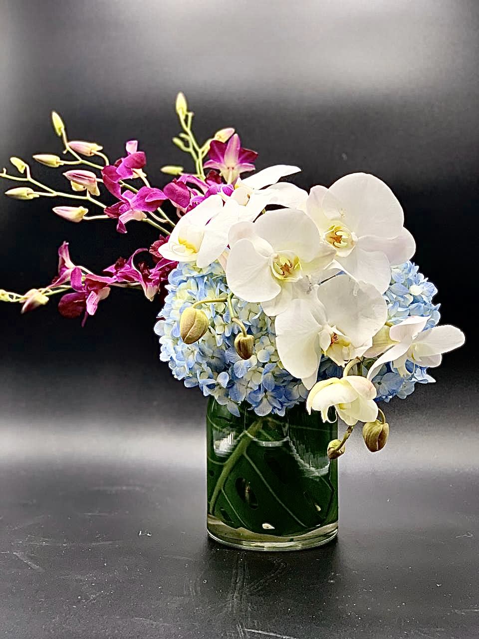 White orchids and blue hydrangeas in a glass vase with magenta orchid sprays