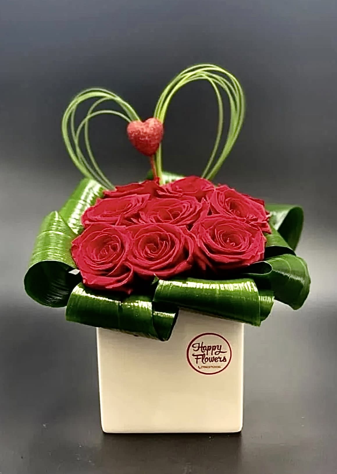 Red roses in a white box with green ribbon loops and a heart topper