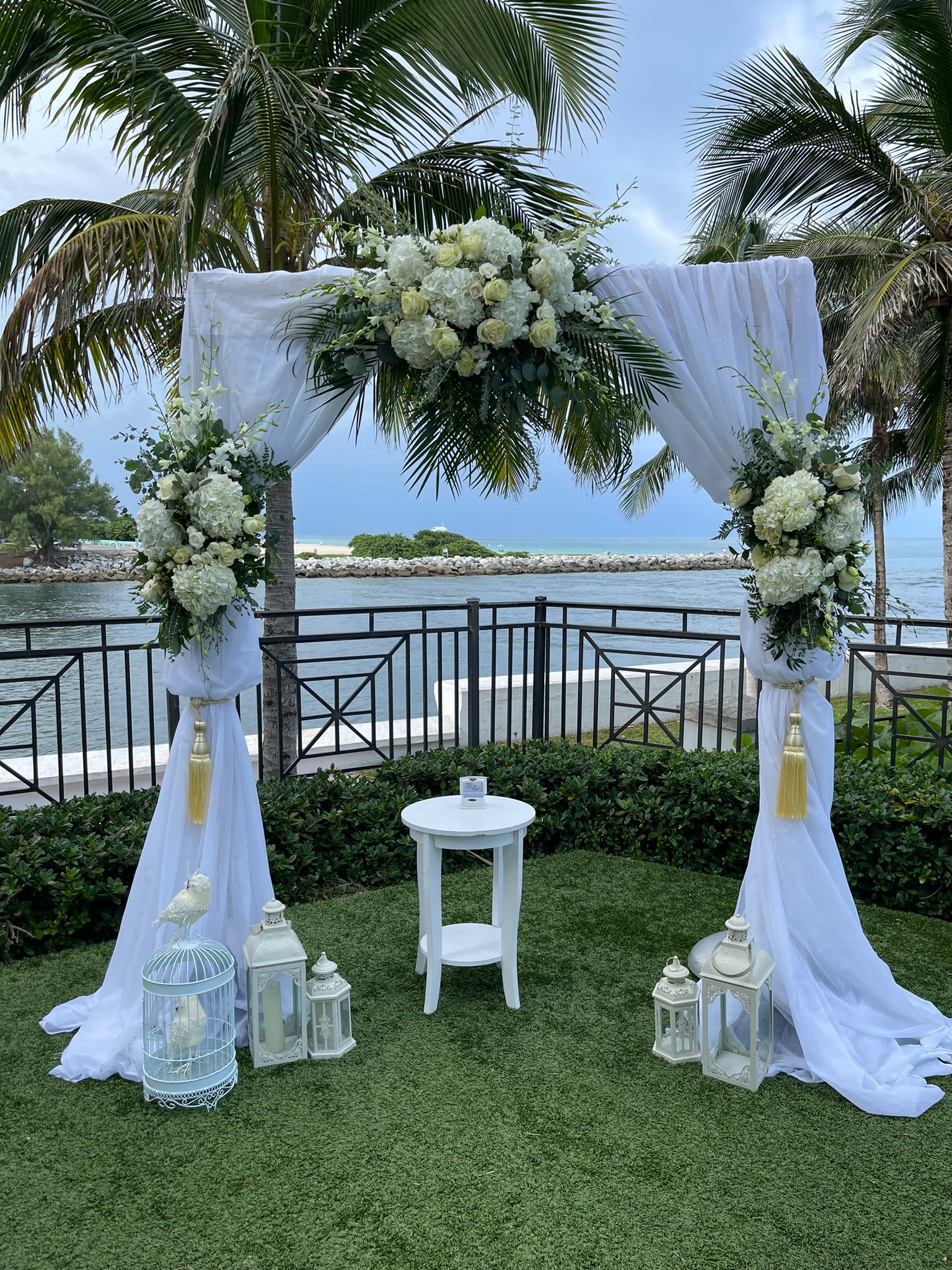 White floral wedding arch with draped fabric and lanterns by the water