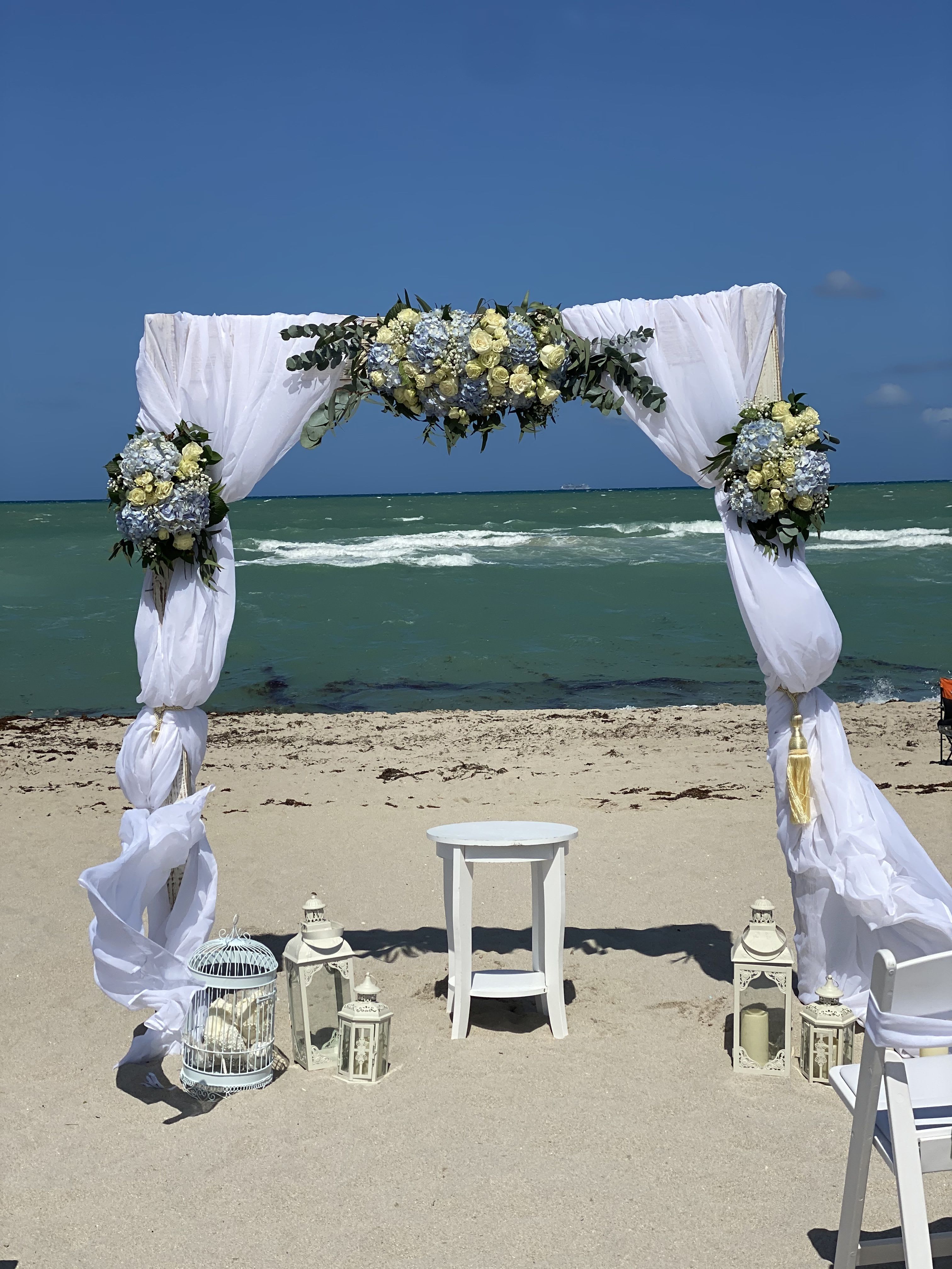 Beach wedding arch with white drapes and blue-and-cream floral arrangements