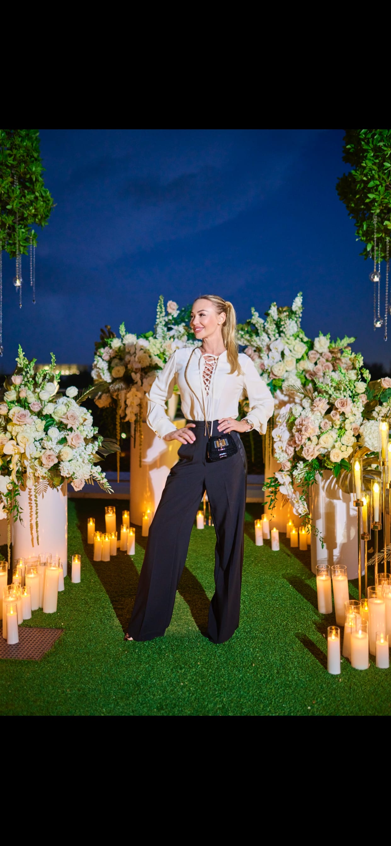 Woman posing between large floral arrangements and candles at night