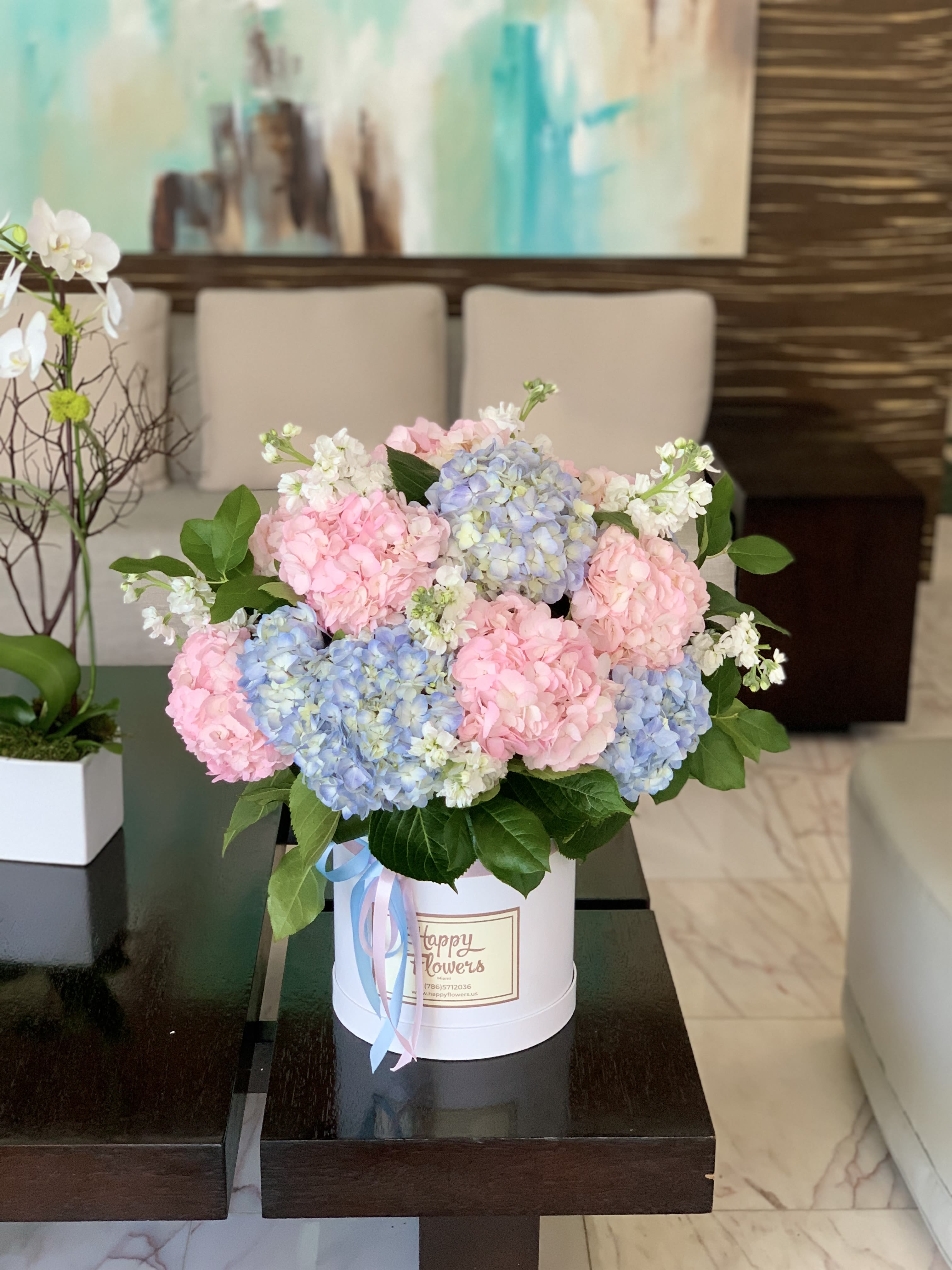 Maria - A lush cluster of pastel hydrangeas in pink, blue, and pale lavender fills a white round hatbox for a romantic birthday bouquet or springtime thank-you gift. The airy, celebratory color palette and full, rounded silhouette make this flower arrangement perfect for anniversaries, Mother's Day, or any moment you want the person you're thinking of to feel truly seen and celebrated.  Dense hydrangea heads are softened with small white filler blooms and glossy green foliage for rich texture and gentle movement. The presentation in a white hatbox tied with soft blue and pink ribbon gives a polished, gift-ready look that arrives ready to display on a table or desk.  This arrangement reads as both elegant and effortless-ideal for someone who loves soft pastels and classic styling. Size and care: keeps best in a cool spot away from direct sun; refresh water daily if transferring to a vase, and handle the hatbox gently when moving.