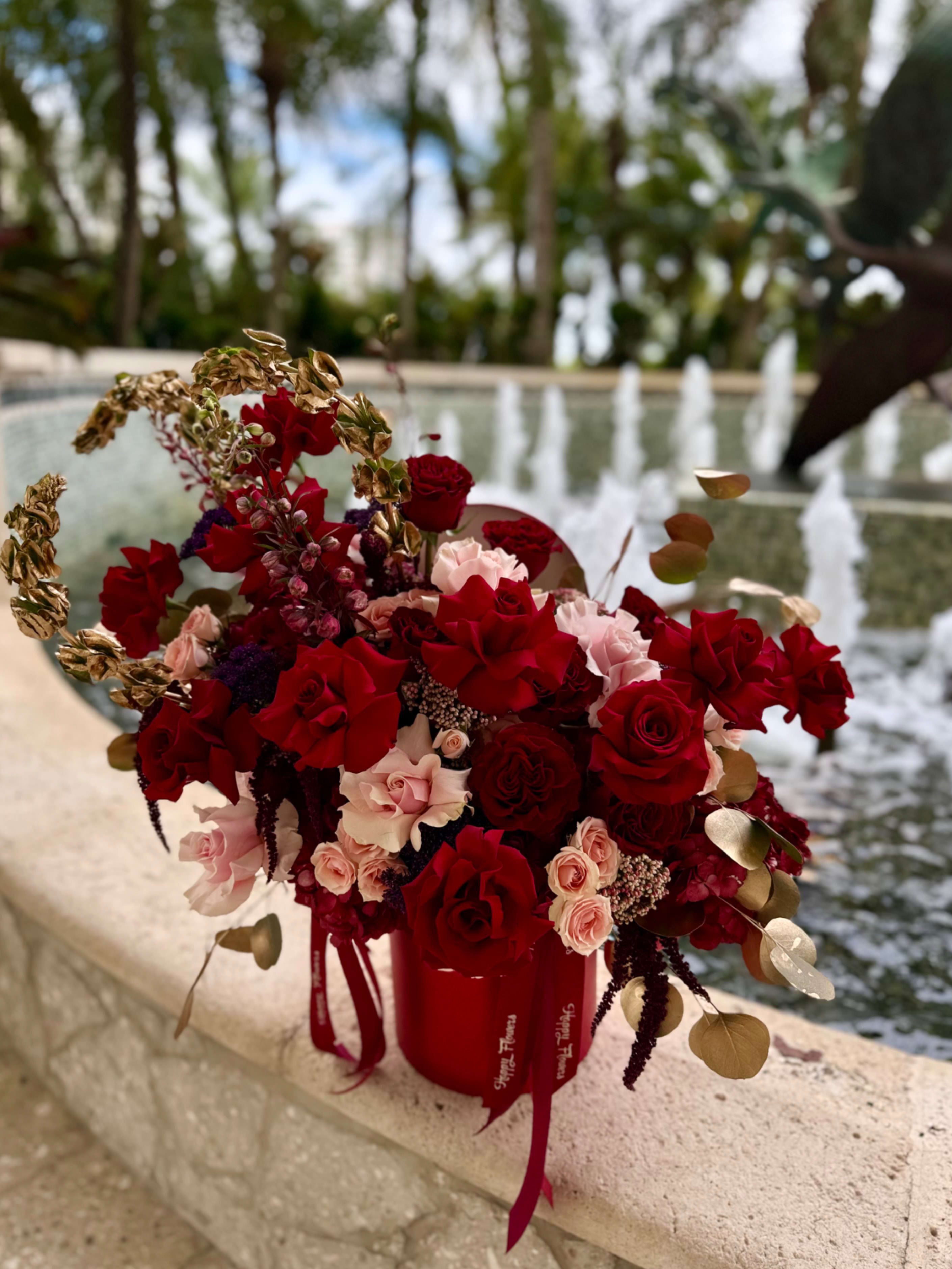 Red passion - A lush cluster of red and blush roses-accented with spray roses and deep purple filler blooms-creates an immediate feeling of passion and celebration, perfect for a romantic bouquet or anniversary arrangement. Set in a red cylindrical box with elegant ribbon tails, this flower arrangement reads as a gift-ready statement that will make the person you're thinking of feel deeply cherished.  Gold-toned decorative foliage arcs above and around the blooms, adding a dramatic, celebratory contrast that elevates the rich red palette into something luxurious and formal. The mix of full roses, smaller spray roses, and dark accent flowers gives the arrangement depth and texture while the red container and ribbon finish the look with polished presentation.  Ideal for Valentine's Day, anniversaries, or any occasion where you want to express love and admiration, this arrangement arrives ready to display-no extra vase needed. Its dense, compact design keeps blooms looking abundant; to prolong freshness, keep the box in a cool spot away from direct sun and replace water per the included care instructions.