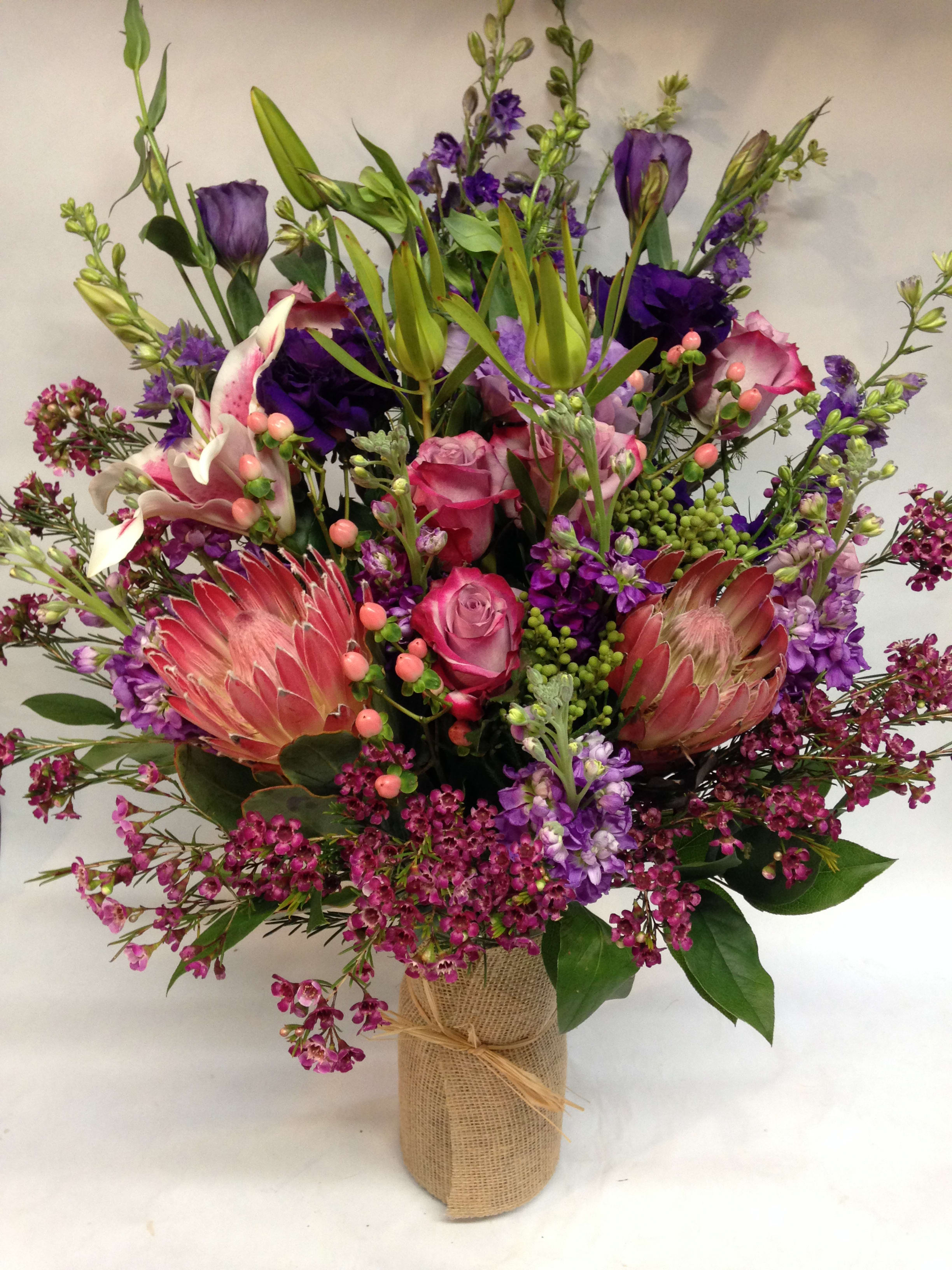 Royal Arrangment - This large arrangement comes complete with king proteas, lisianthus, deep purple roses, stargazer lilies, wax flower, safari branches, pink hypericum, purple larkspur and stock. (Some flowers may be subject to change depending on the season. We will contact you with available flower substitutions; please contact us with specific questions if you are concerned with flower availability, thanks!)  FEATURED ARRANGEMENT: 450.00 (REGULAR PRICE)