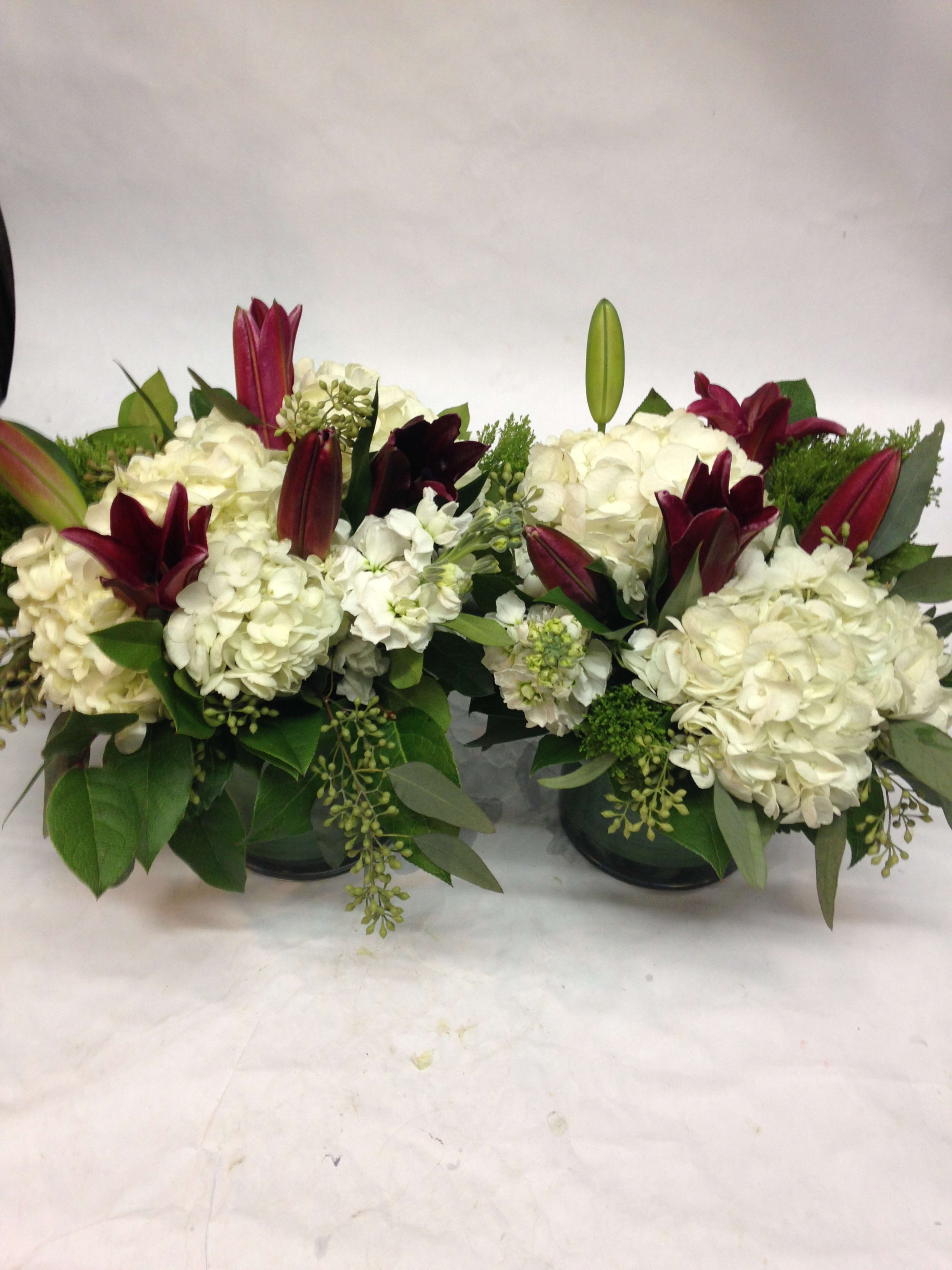 Two white floral arrangements with burgundy lilies in dark containers