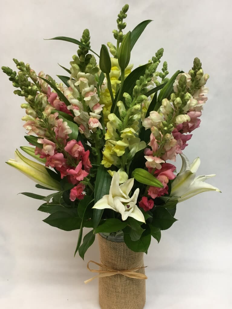 Tall bouquet of pink, yellow, and white snapdragons with white lilies in a burlap-wrapped vase